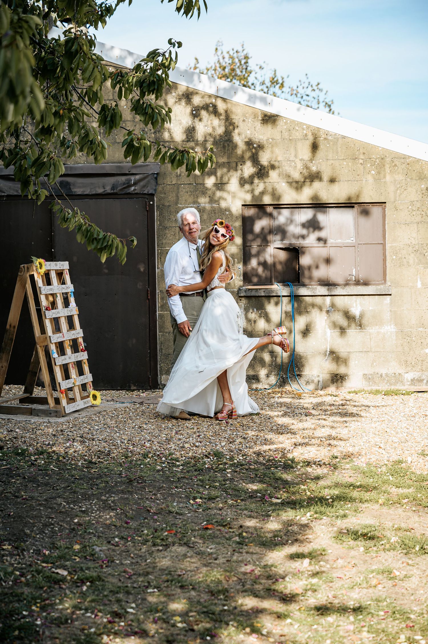 Bride in white two-piece gown poses playfully with older gentleman outside rustic building in sunlight.