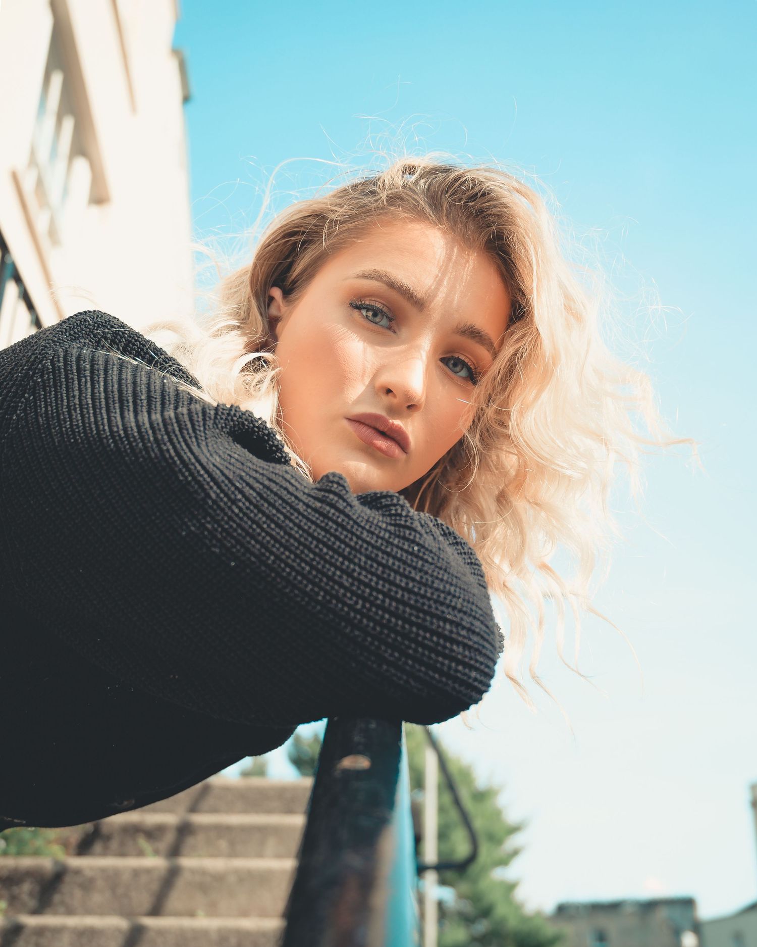 Blonde woman in black knit sweater leans on railing under blue sky, looking up at camera.