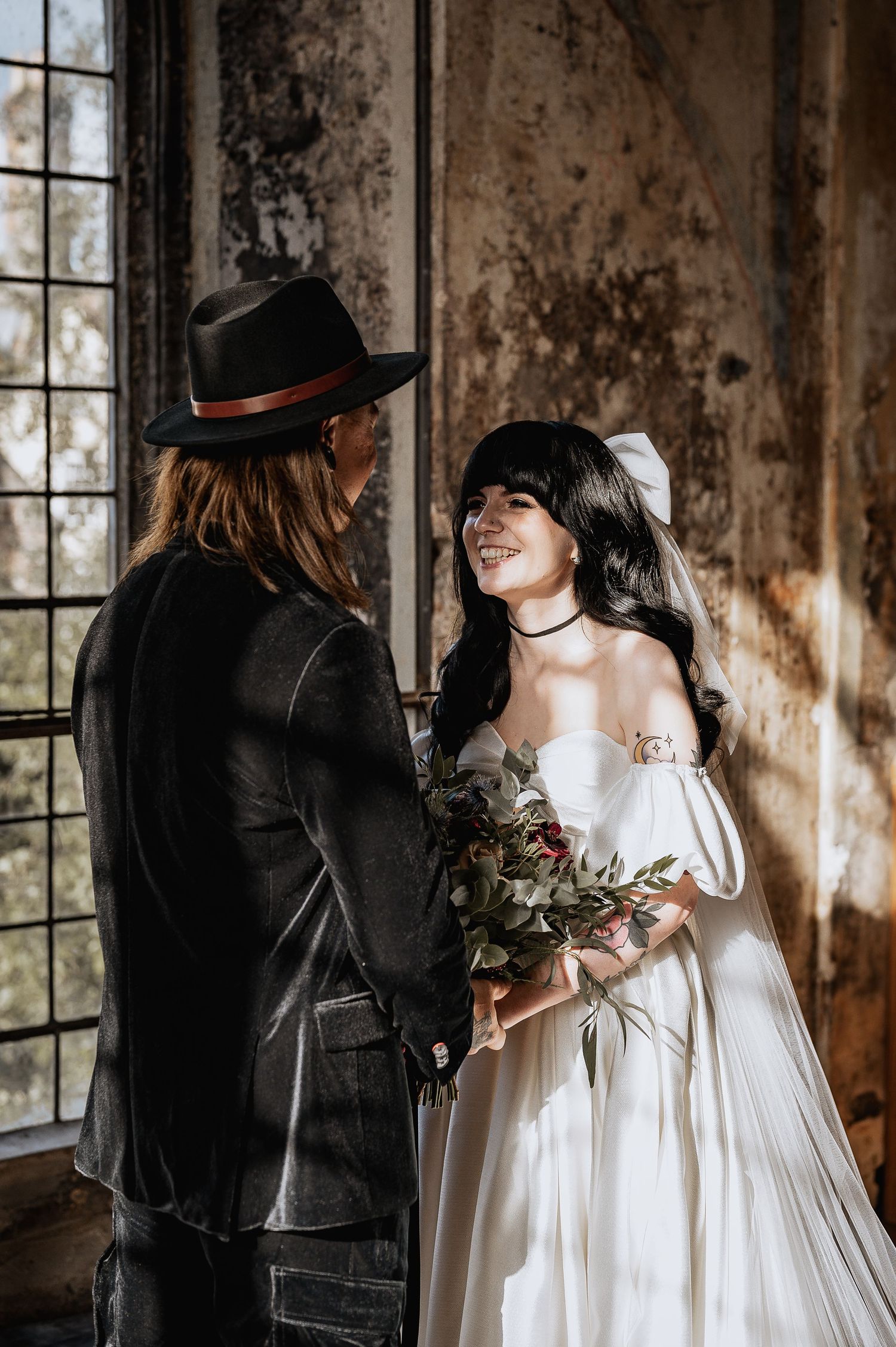 Bride in white gown with bouquet smiling at groom in black hat by window.