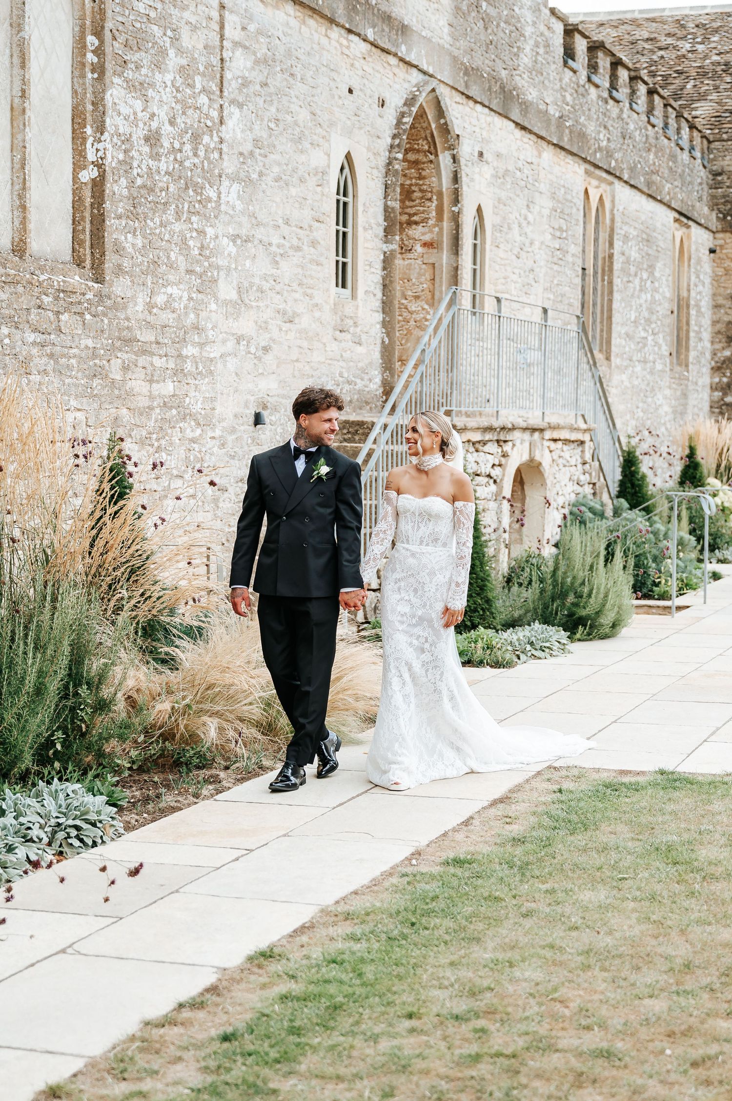 Bride in off-shoulder lace gown and groom in black tuxedo walk hand in hand by stone church.