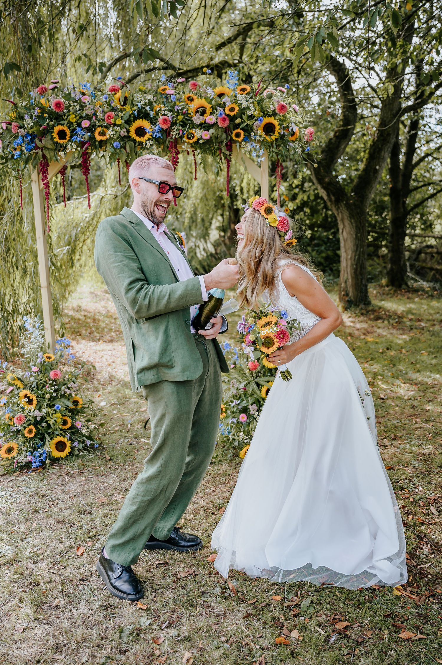 Smiling groom in green suit opens champagne bottle with bride in floral crown under sunflower arch.