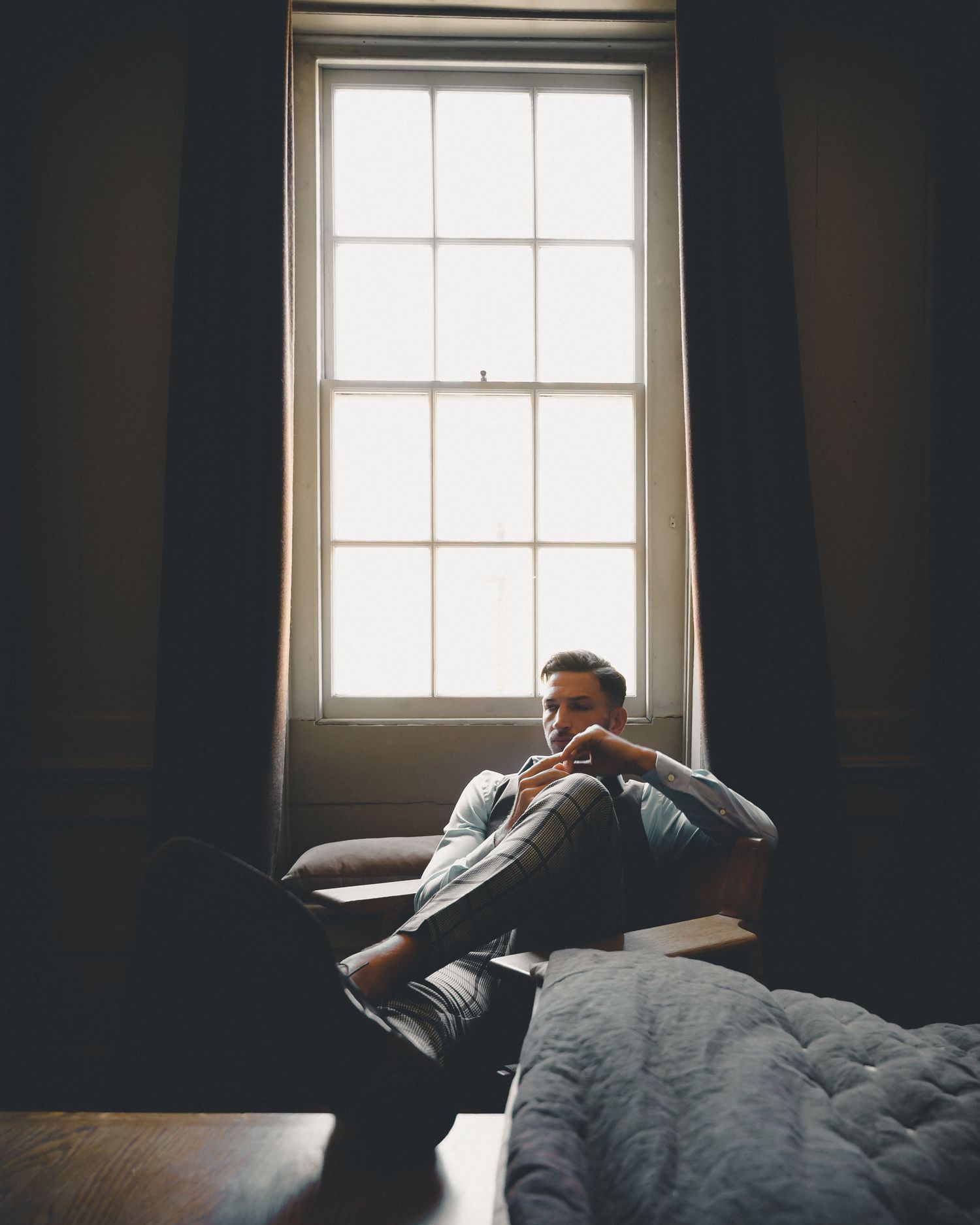 Pensive man sitting by large window in dimly lit room with natural light backdrop.