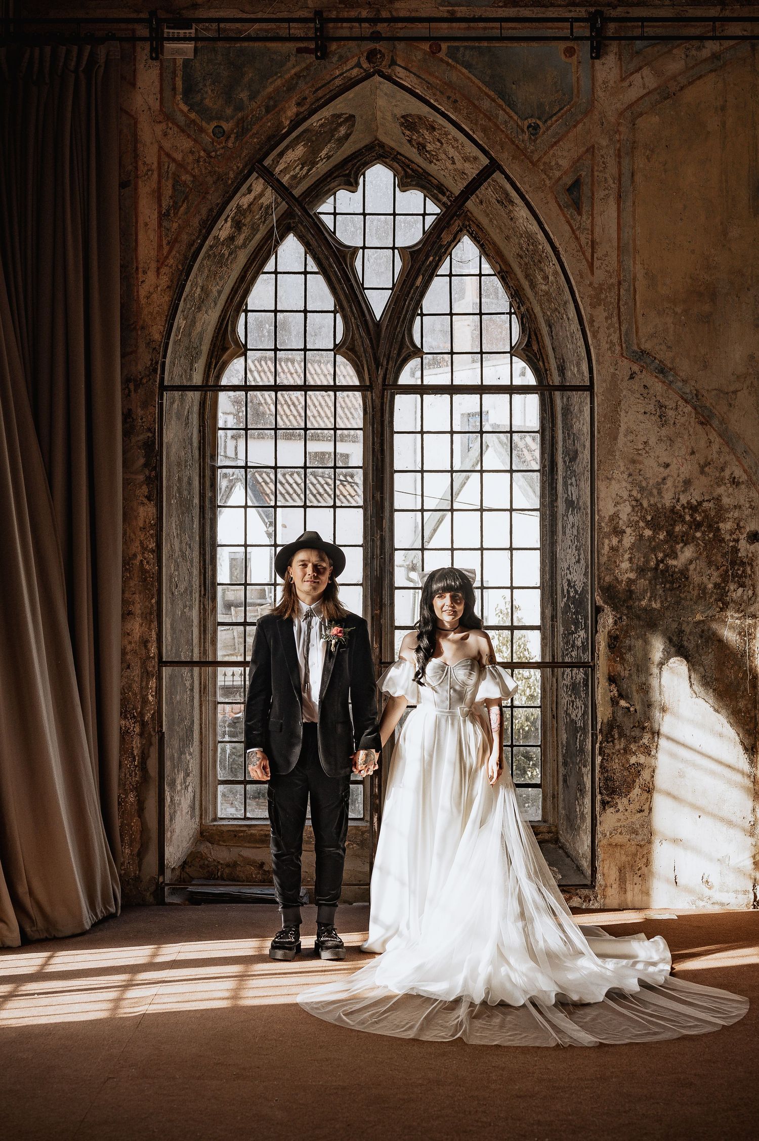 Bride and groom standing before grand gothic arched window in sunlit rustic venue.