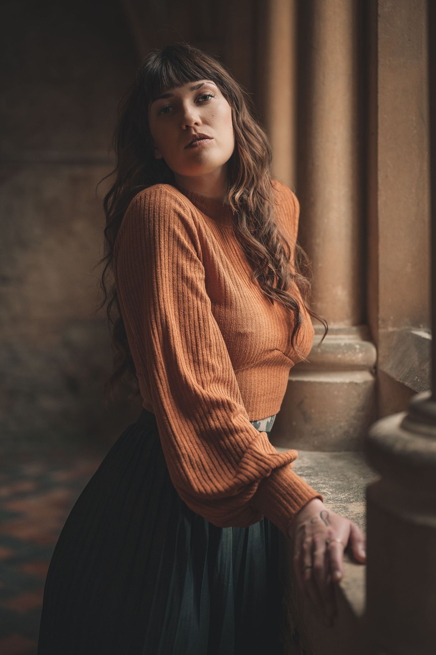 Young woman in rust orange ribbed sweater leaning against stone column in elegant archway.