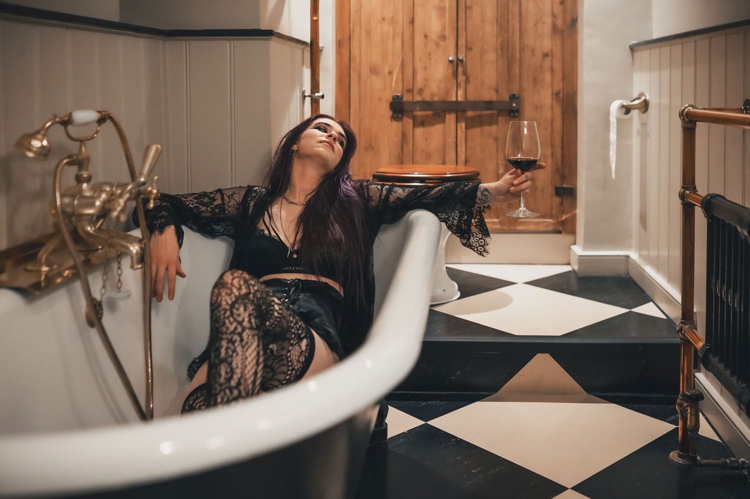 Woman in black lace outfit reclines beside a vintage clawfoot bathtub holding a glass of red wine.