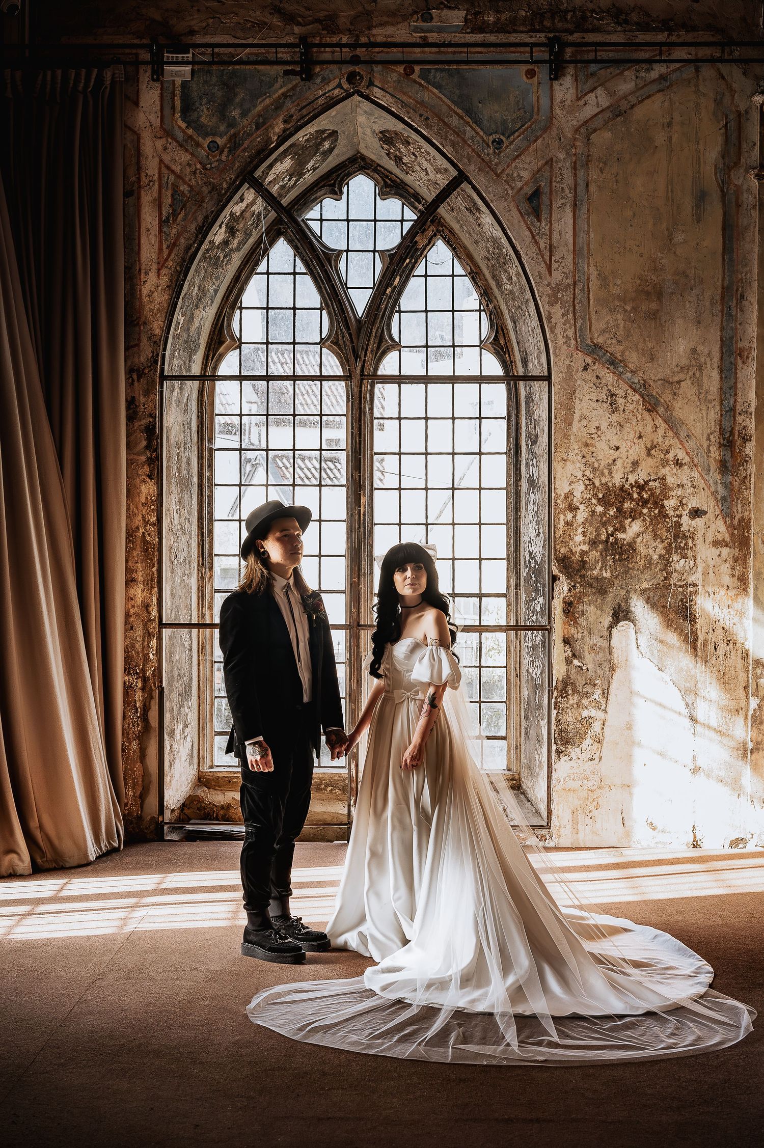 Bride and groom share a romantic moment before a Gothic arched window in a historic venue.