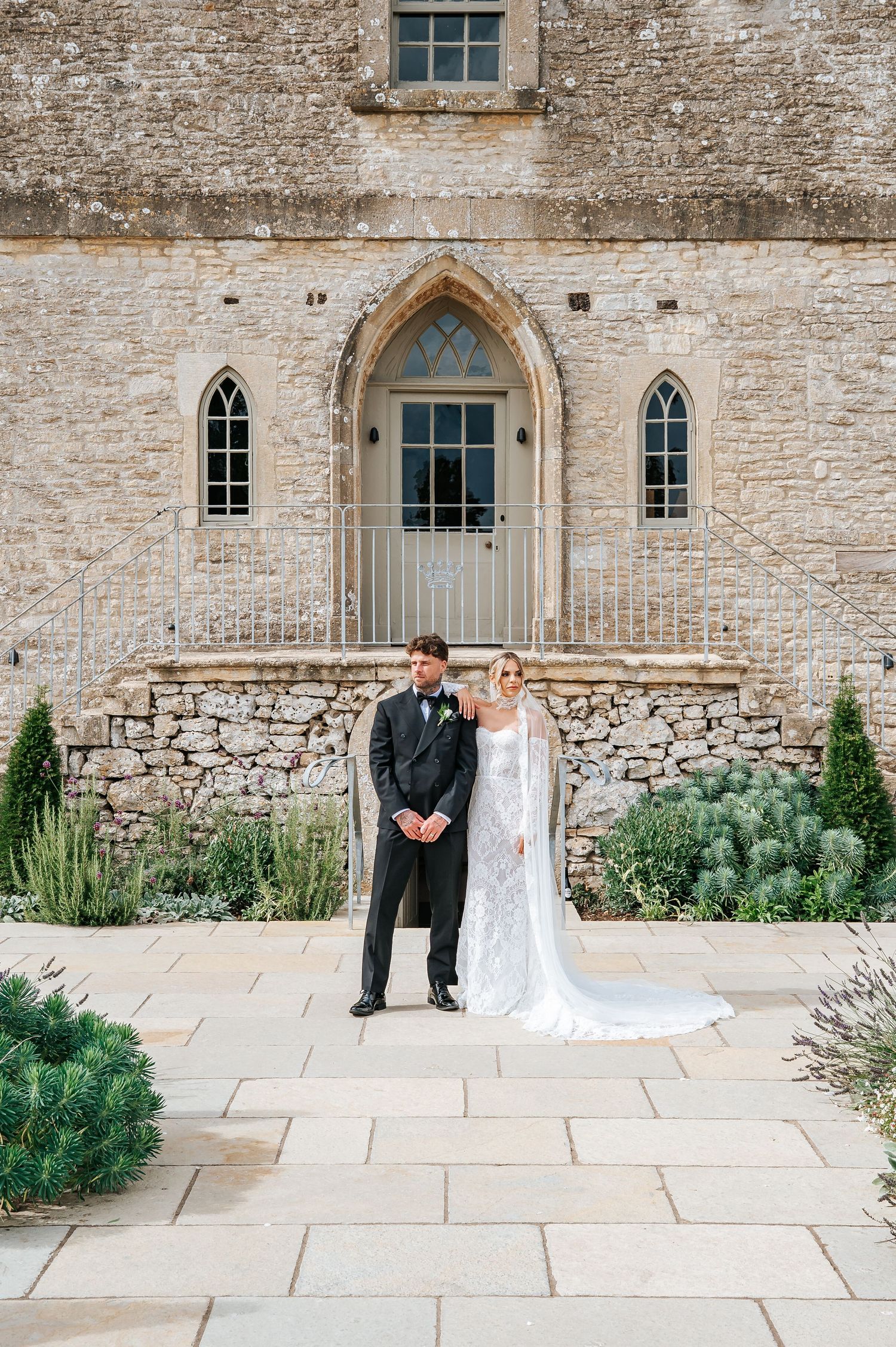 Bride in elegant lace gown and groom in black tuxedo pose before a historic stone church.