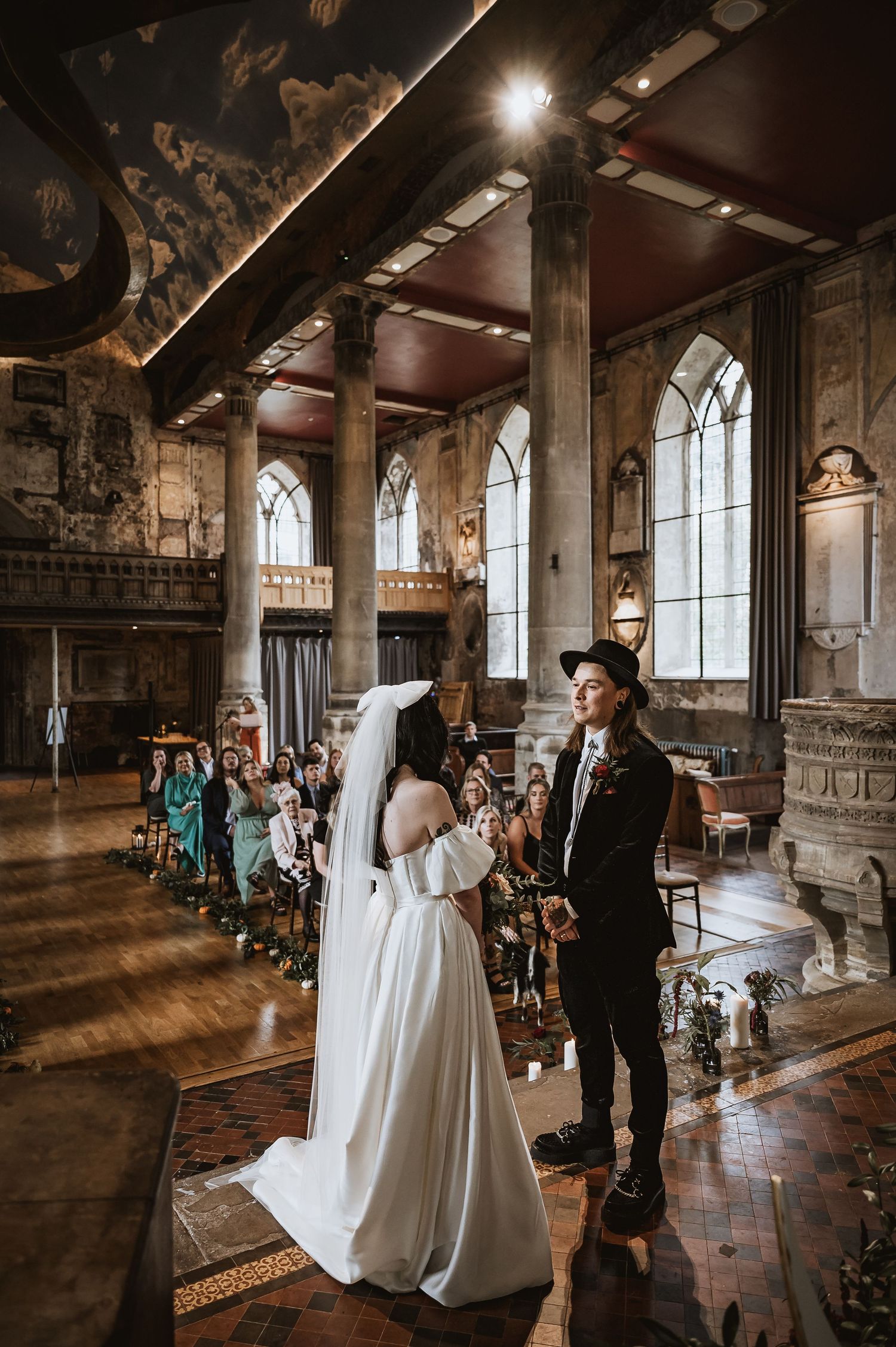 Bride and groom exchanging vows in ornate gothic hall with painted ceiling and arched windows.