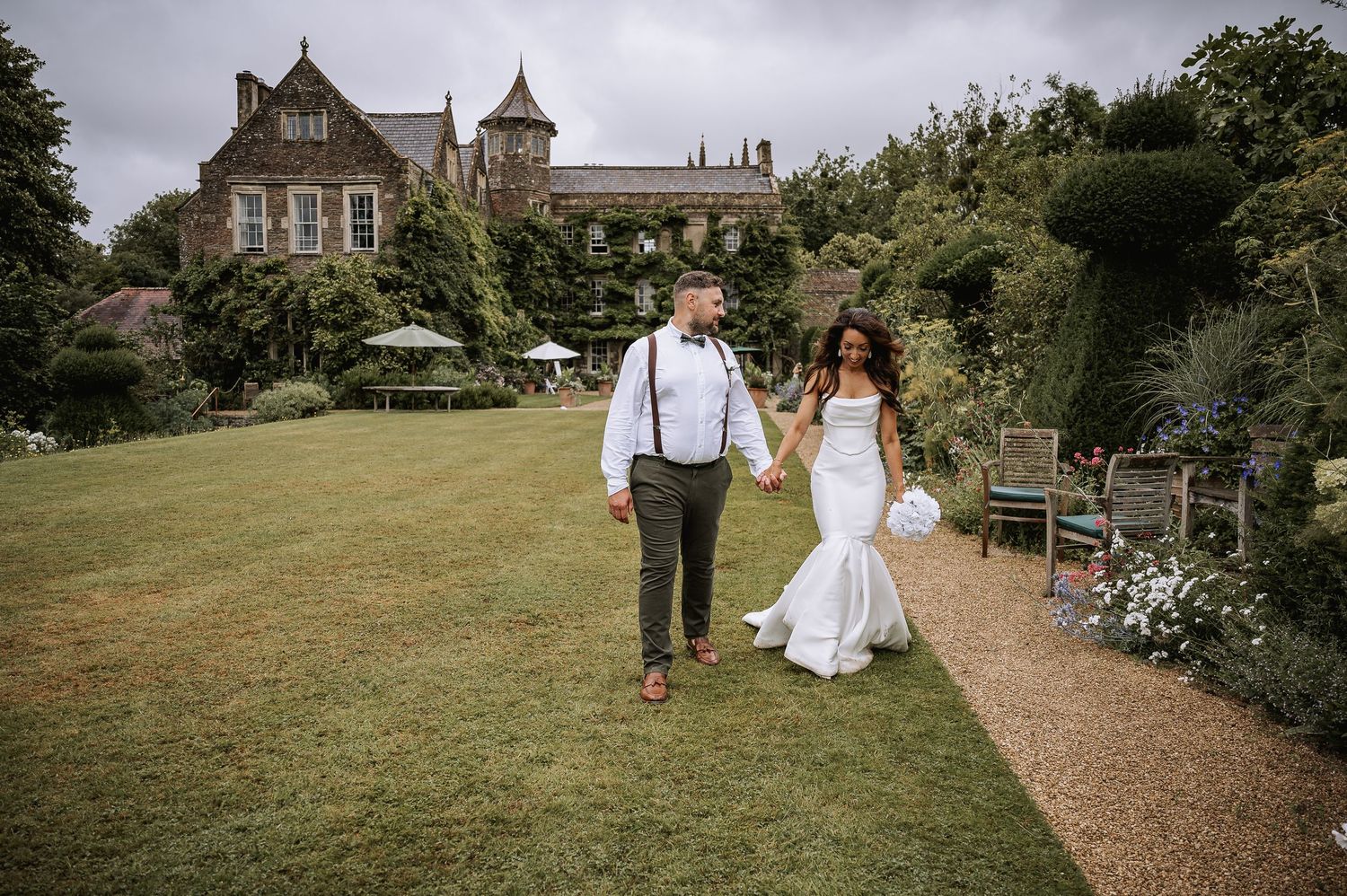 Bride and groom walking hand in hand through lush garden with historic manor house backdrop.