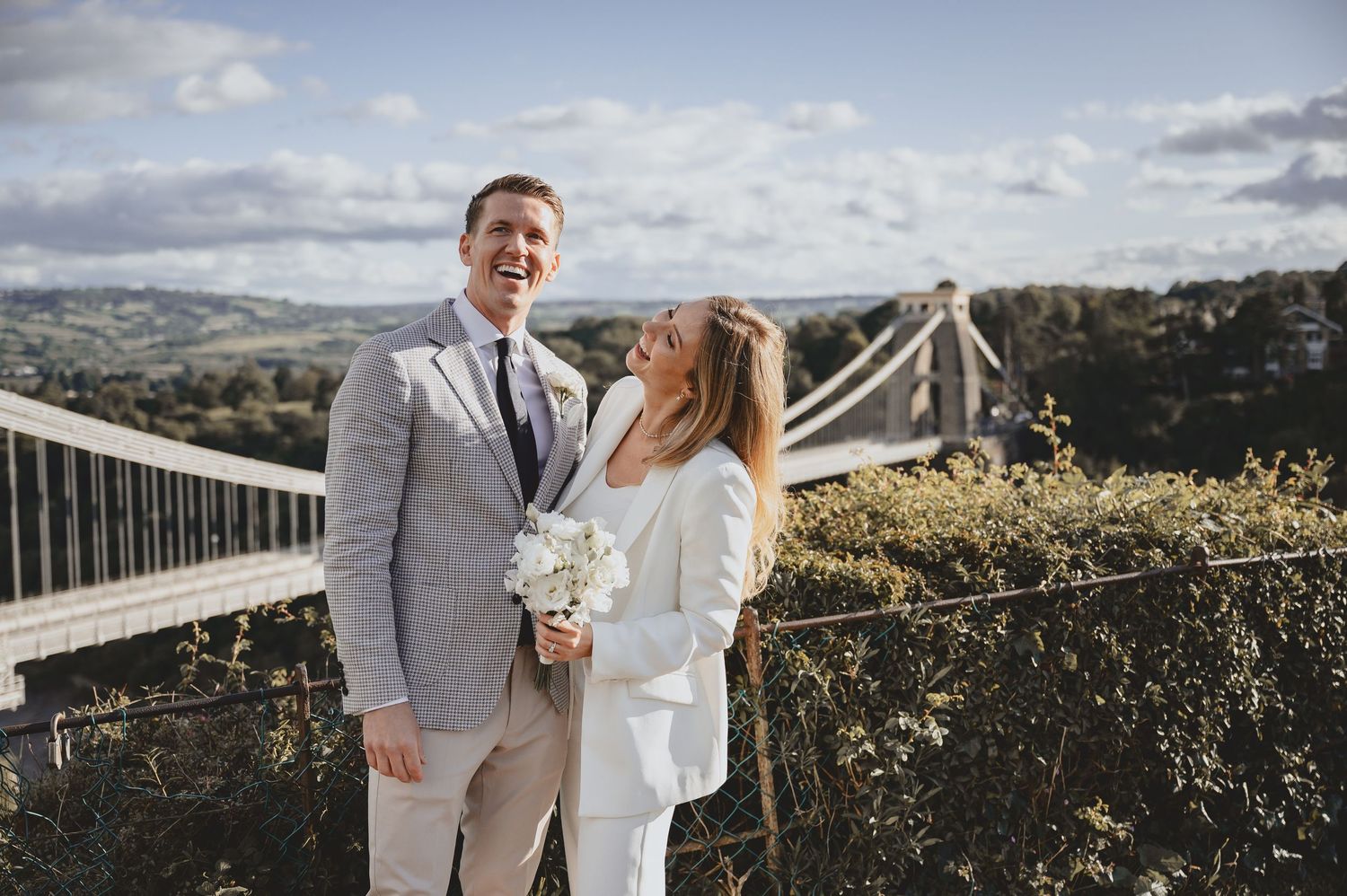 Smiling couple in wedding attire posing outdoors with scenic countryside and suspension bridge in background.