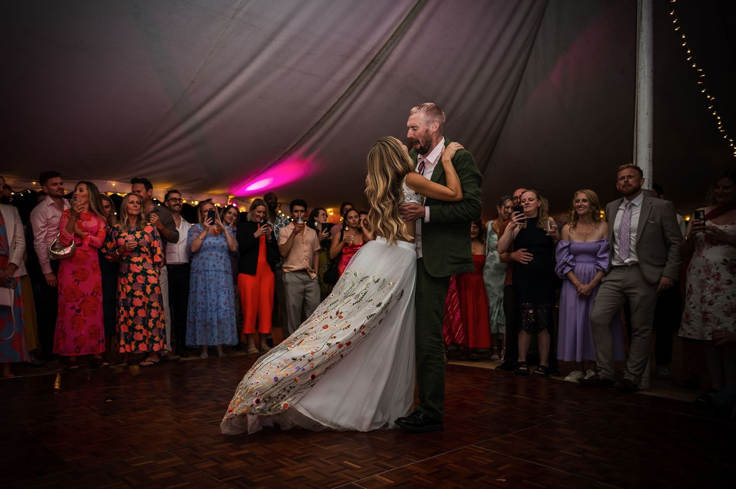 Bride and groom share romantic first dance surrounded by guests inside elegant wedding reception tent.