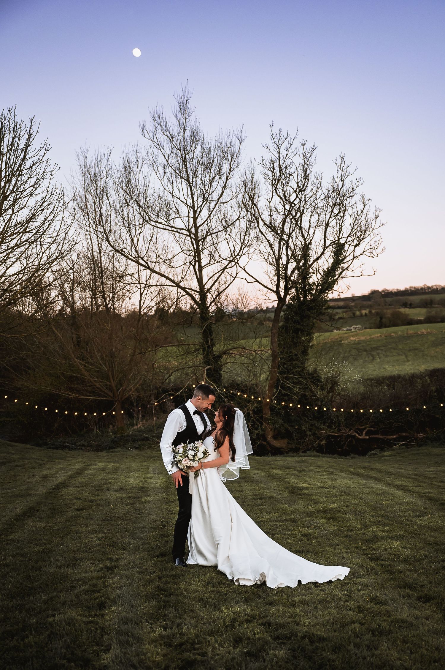 Bride and groom embrace at dusk under a full moon in a scenic countryside wedding setting.