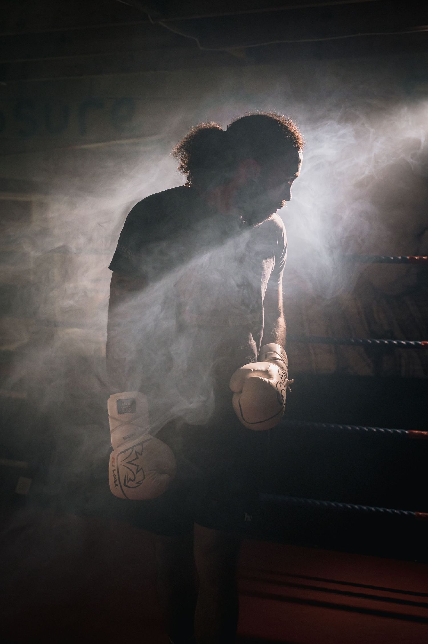 Silhouetted boxer with gloves standing in smoky boxing ring with dramatic backlight.