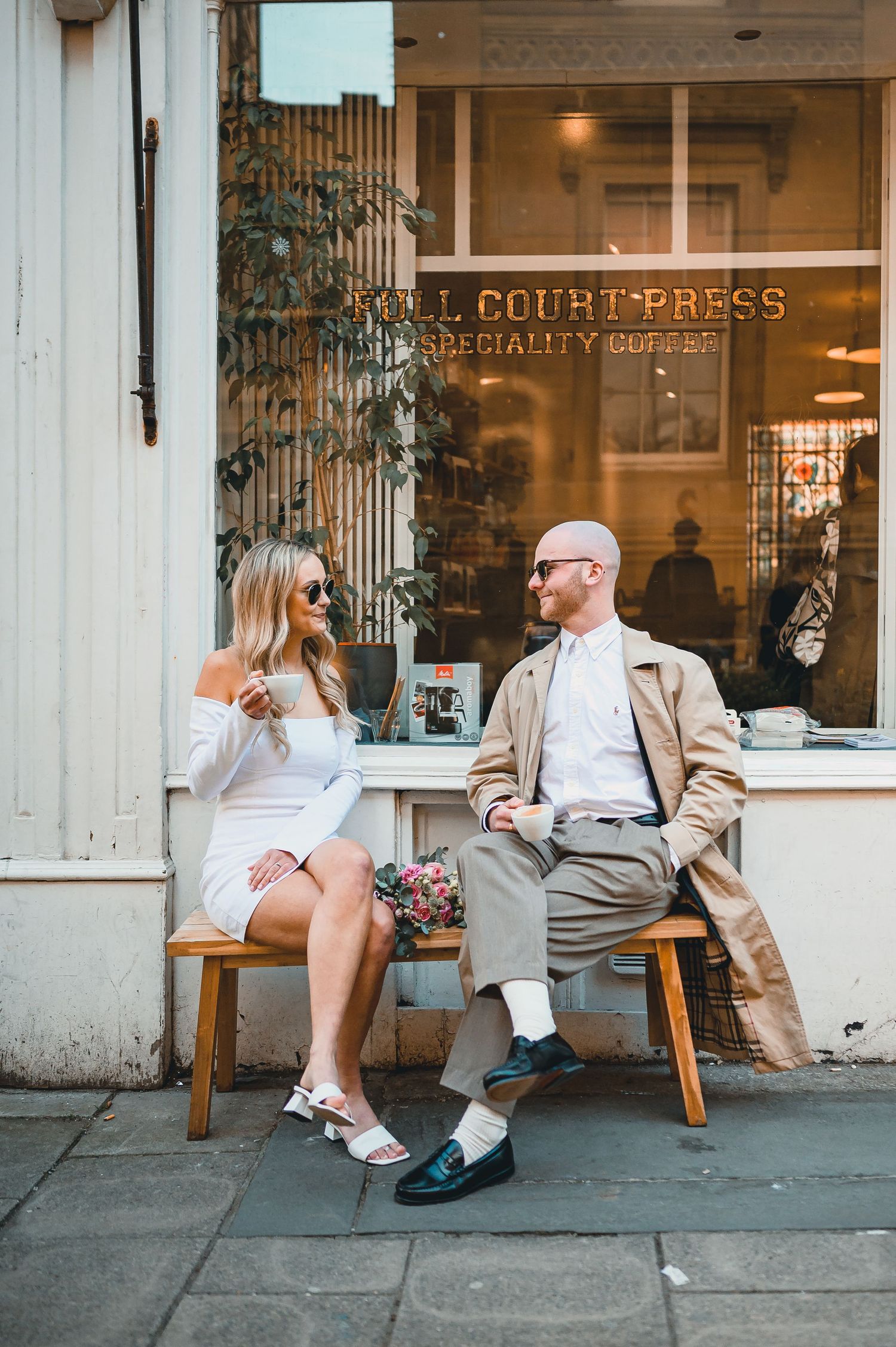 Couple sitting outside Roll Court Press specialty coffee shop, woman in white mini dress holding coffee cup, man in trench coat.