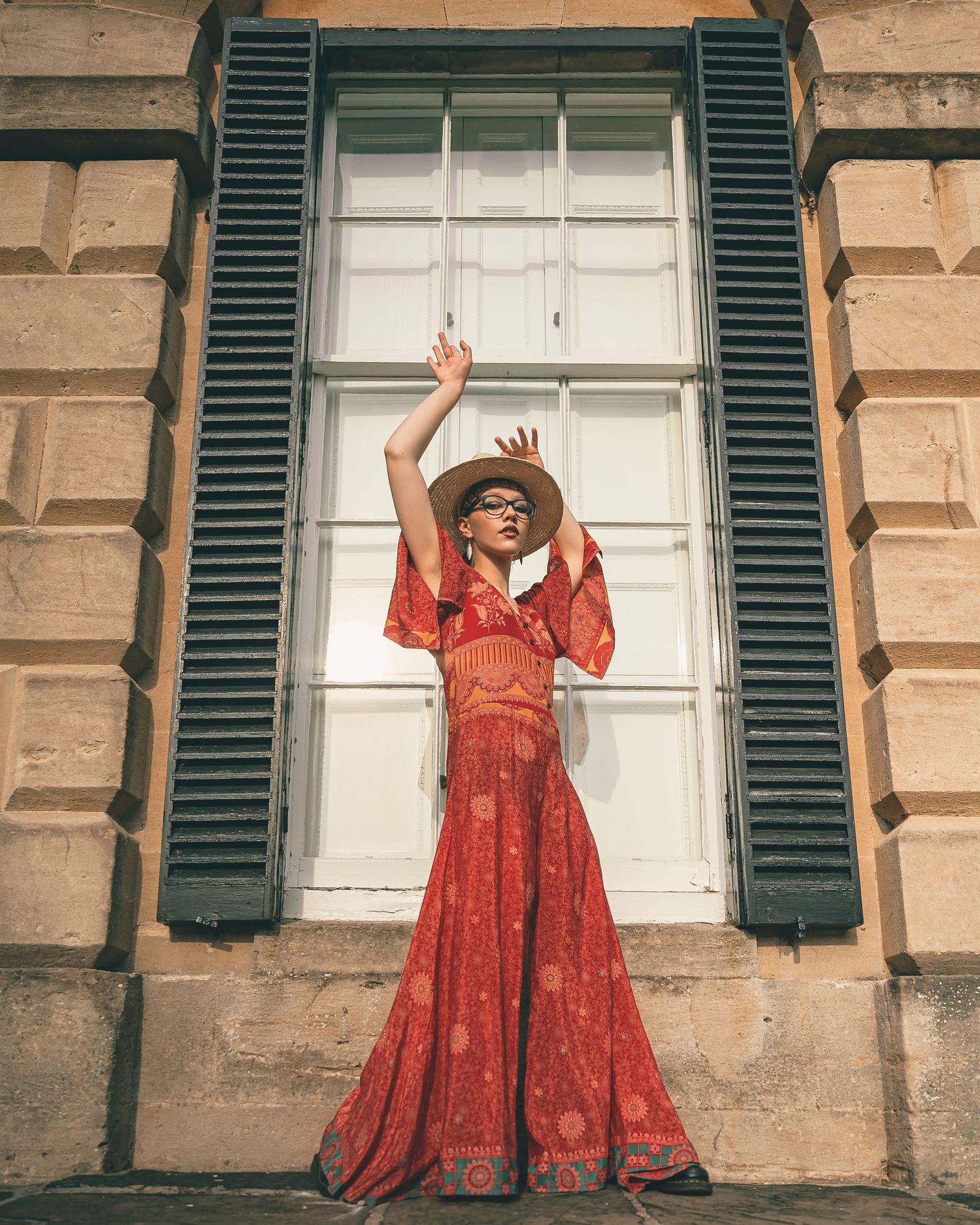 Woman in red boho maxi dress and hat posing with arms raised before a window with dark shutters.