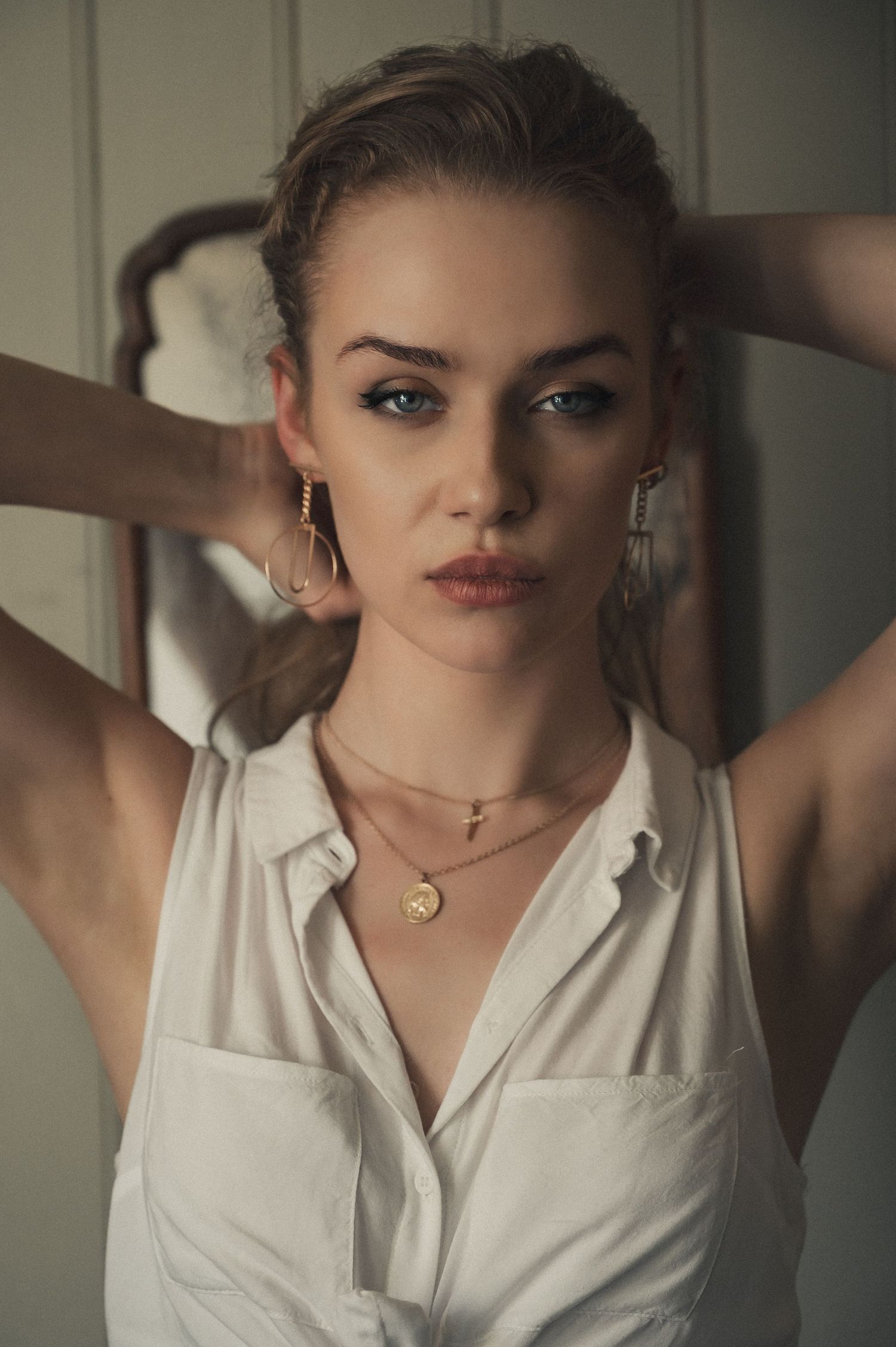 Woman with blue eyes wearing gold hoop earrings and layered gold necklaces in a white sleeveless shirt.