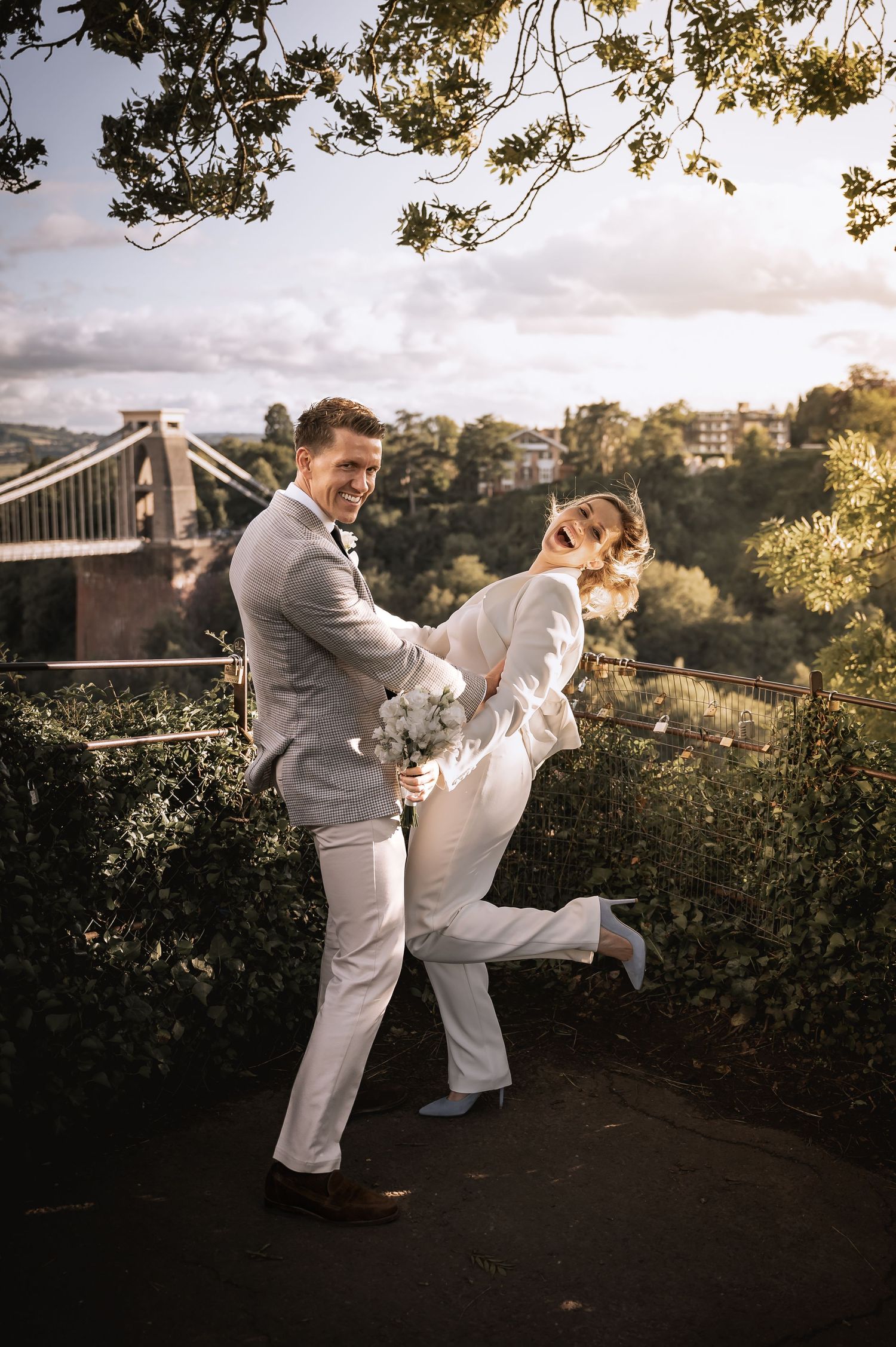 Laughing bride in white pantsuit spun by groom near Clifton Suspension Bridge.