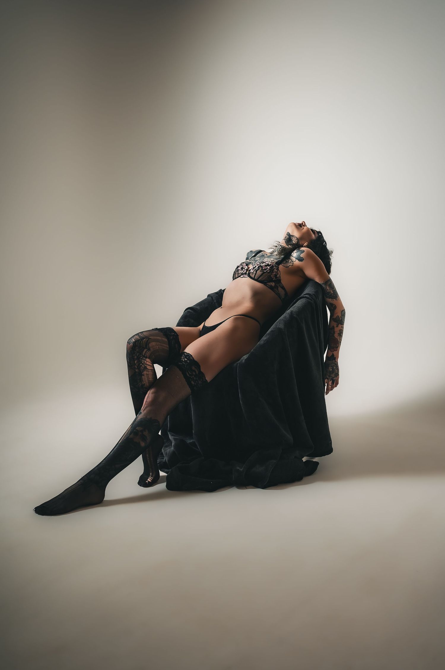 Woman in black lingerie and cape posing dramatically on floor against neutral background.