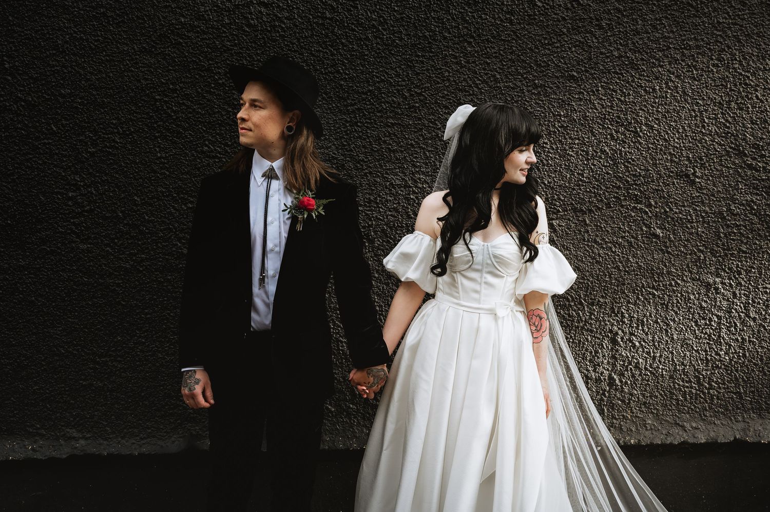 Bride and groom holding hands outdoors, bride in off-shoulder white gown with long veil, groom in black suit.