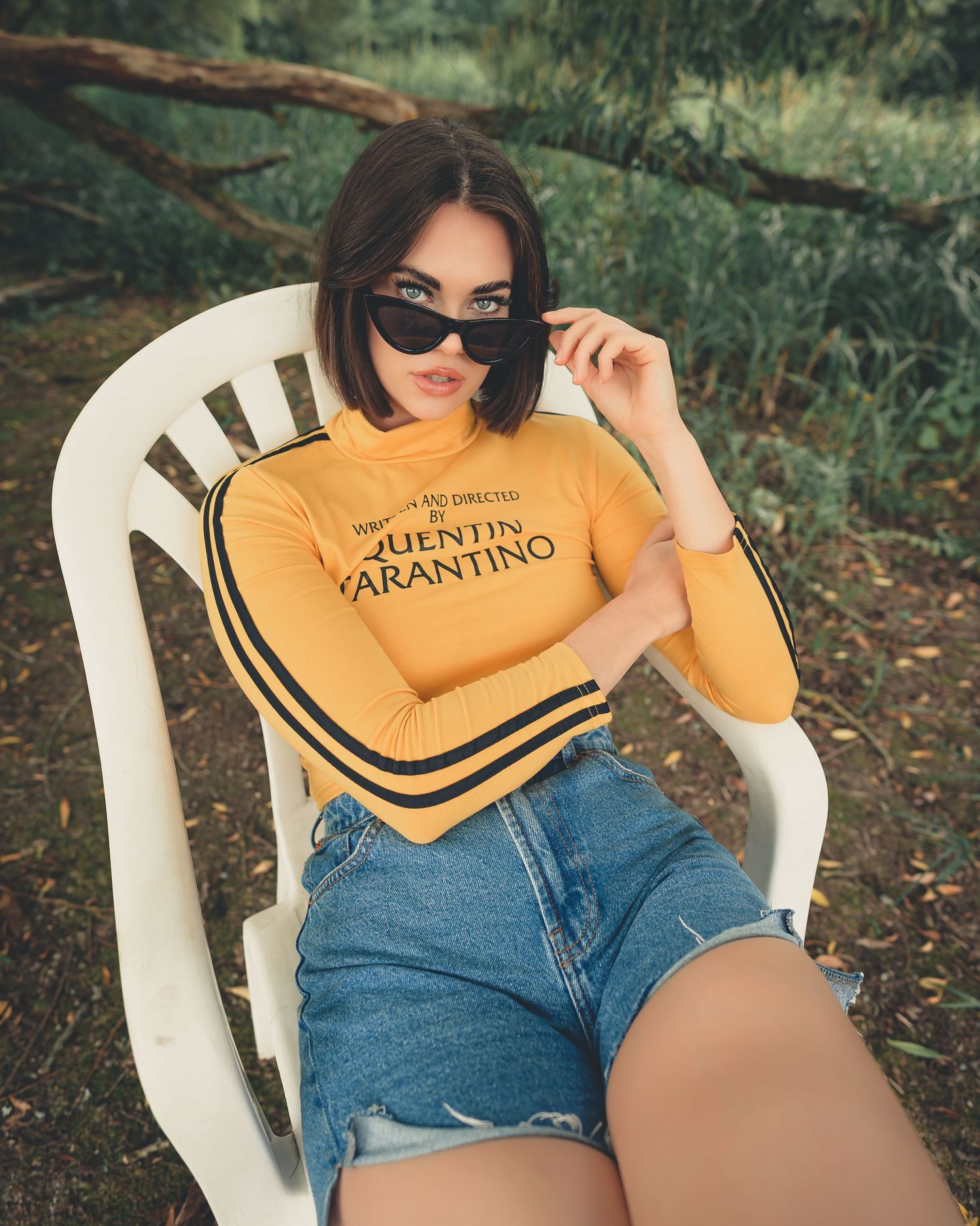 Young woman in yellow Quentin Tarantino long-sleeve top and denim shorts sitting on white chair.