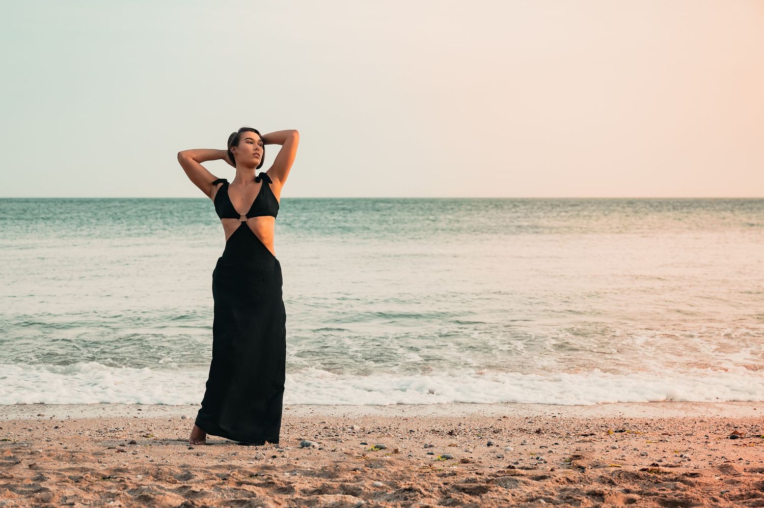 Woman in black cutout maxi dress posing on sandy beach with ocean waves and pastel sky backdrop.