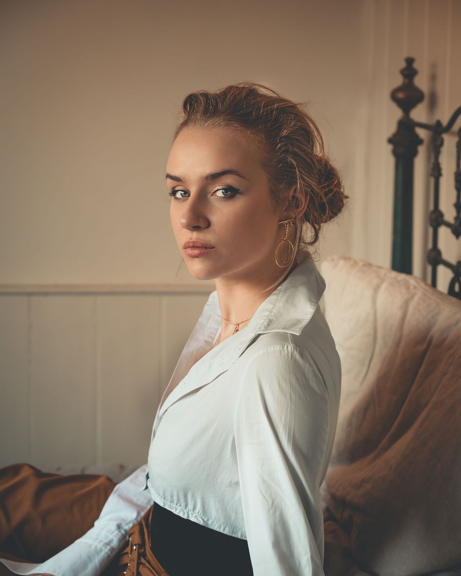 Woman with upswept hair in white blouse and corset sits in warm light beside an antique bed frame.