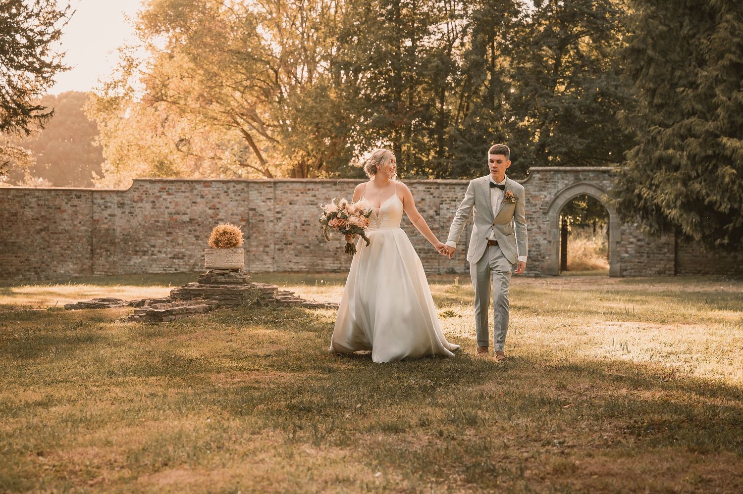 Bride and groom walking hand in hand through golden sunlit garden near historic stone wall.