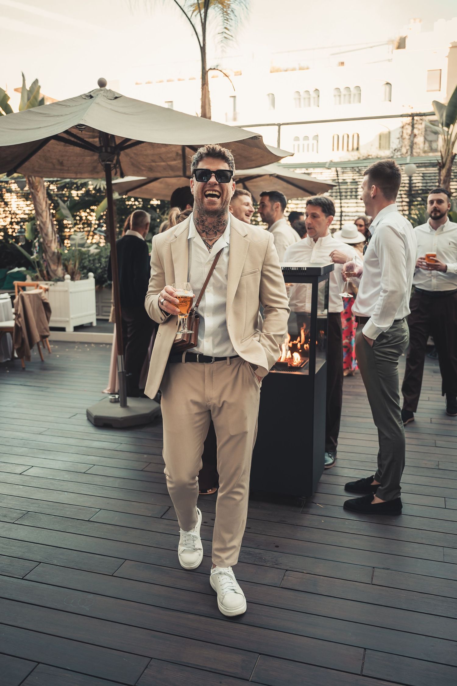 Stylish wedding guest in beige suit holding drink at outdoor rooftop reception.