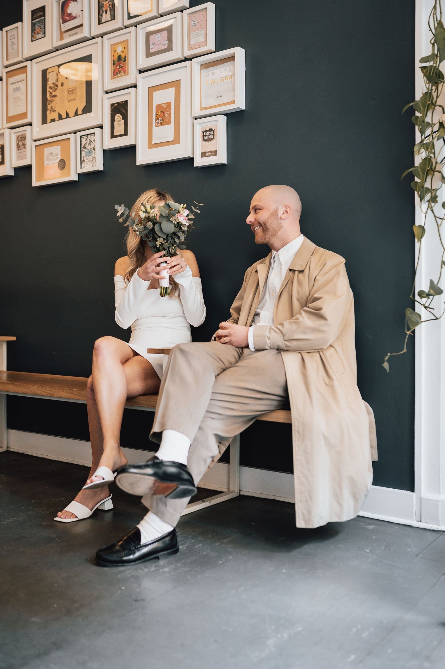 Bride holding floral bouquet and bald groom in beige coat sitting on bench laughing in modern interior.