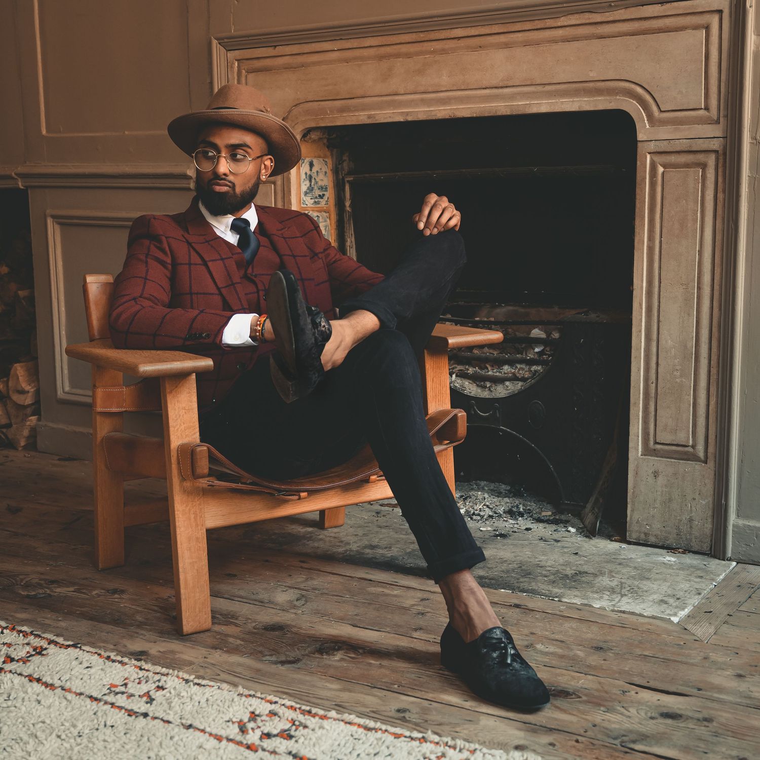 Stylish man in plaid blazer and brown hat sitting in wooden chair by stone fireplace.