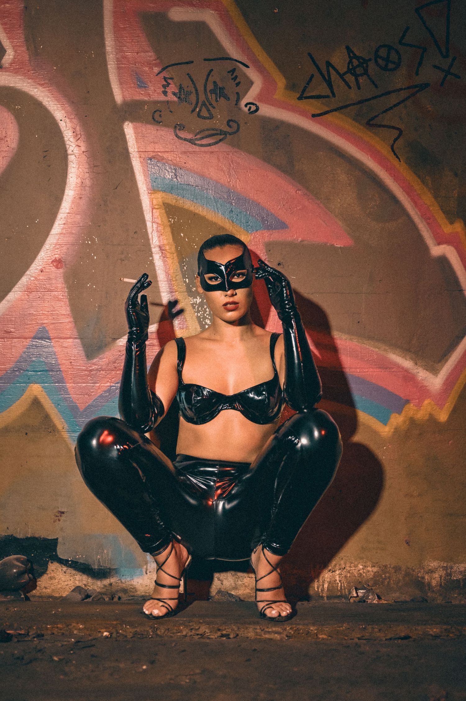 Woman in black latex outfit and masquerade mask posing before colorful graffiti wall.