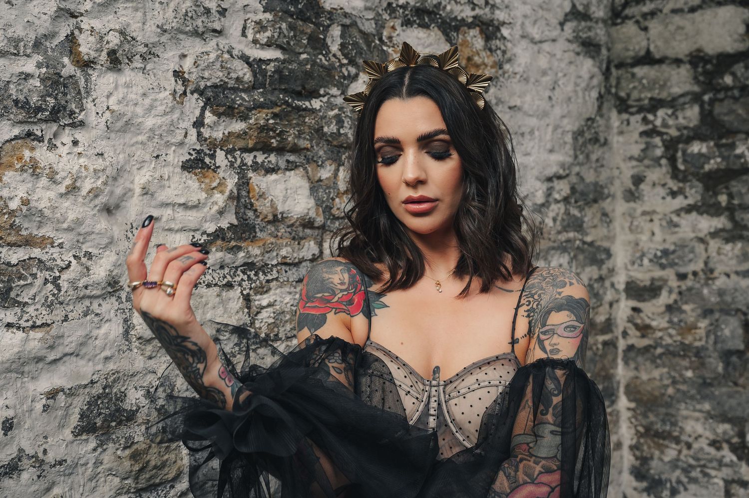Tattooed woman in black lace corset dress with gold crown against stone wall.