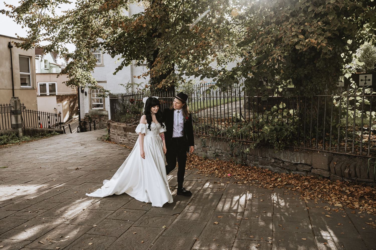 Wedding couple walking hand in hand on cobblestone path surrounded by autumn trees and iron gate.