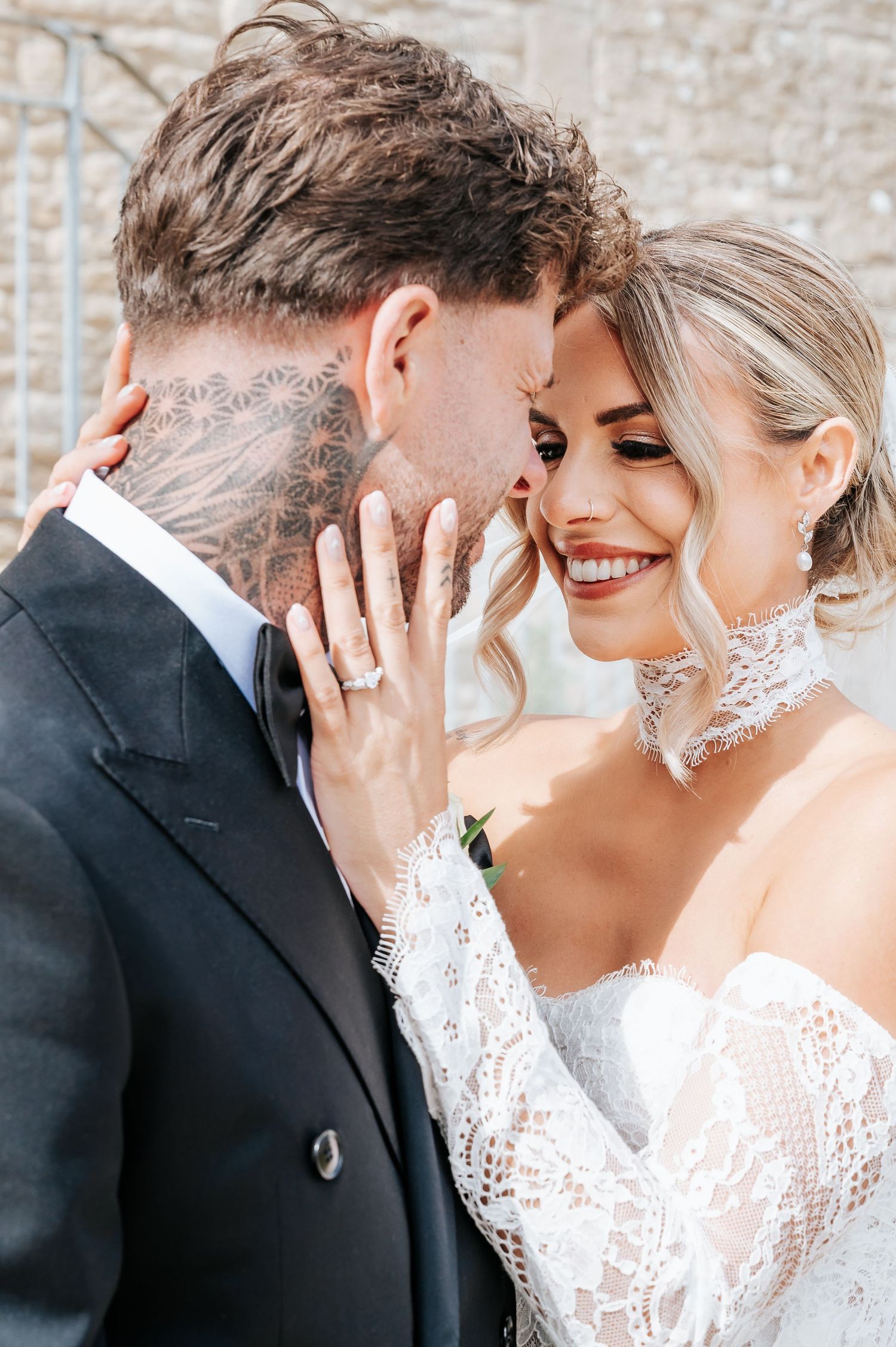 Tattooed groom in black suit and smiling bride in lace choker gown share an intimate moment.