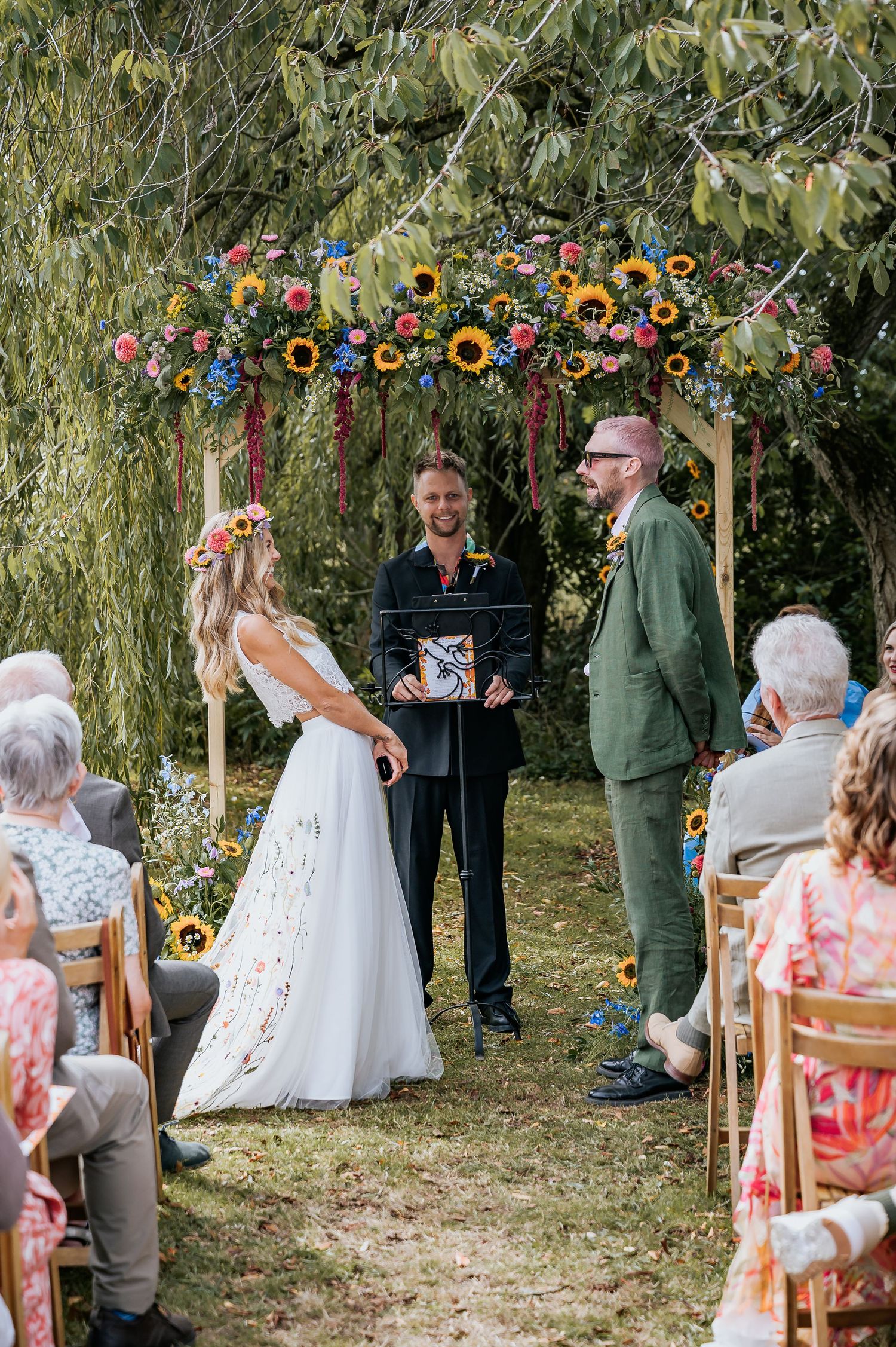 Bride with sunflower crown and groom in green suit exchanging vows under a sunflower-adorned arch.