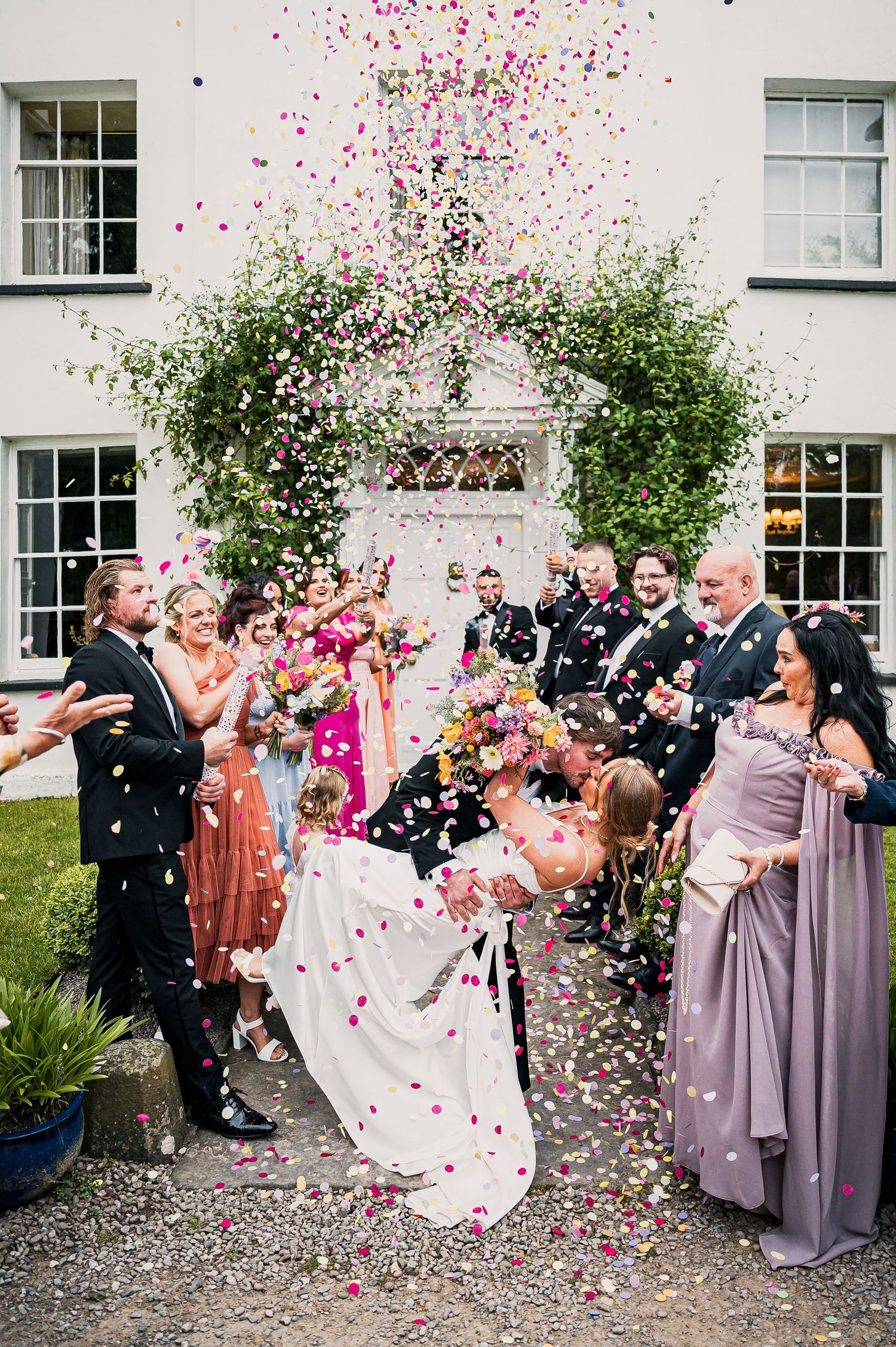 Bride and groom kiss as colorful confetti fills the air surrounded by cheering wedding guests.