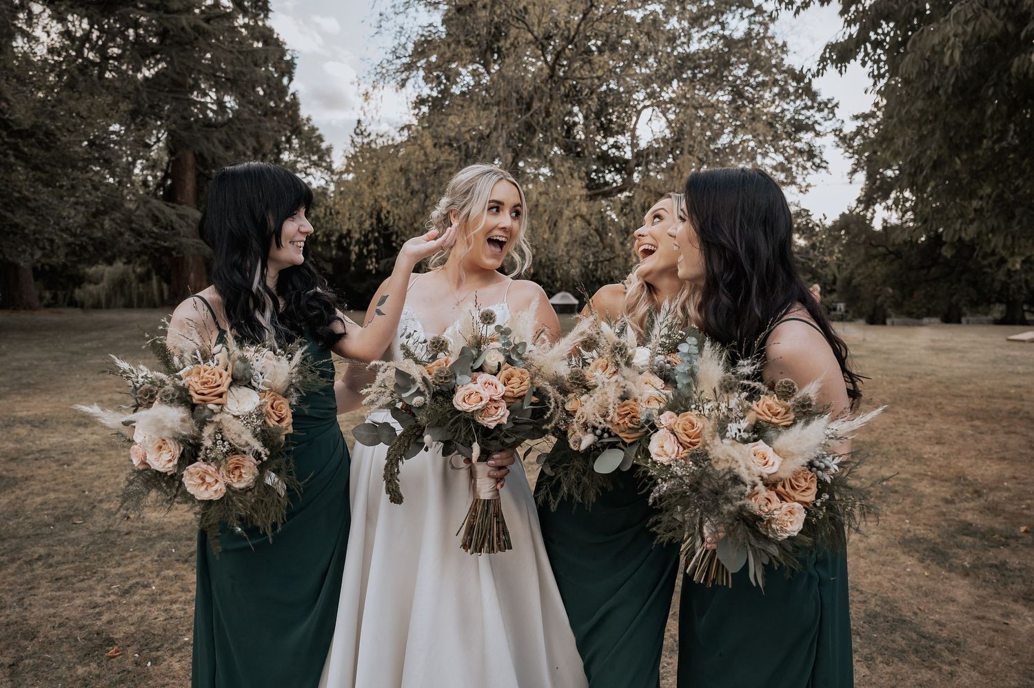 Bride laughing with three bridesmaids in emerald green dresses holding bouquets in park setting.