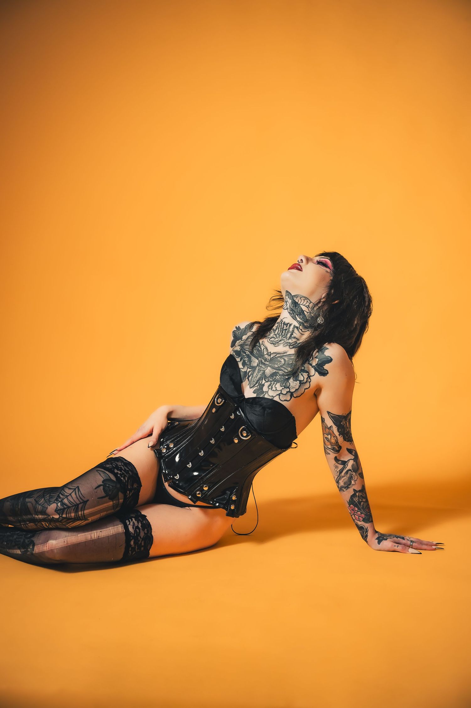 Heavily tattooed woman posing in black corset and stockings against vibrant orange background.
