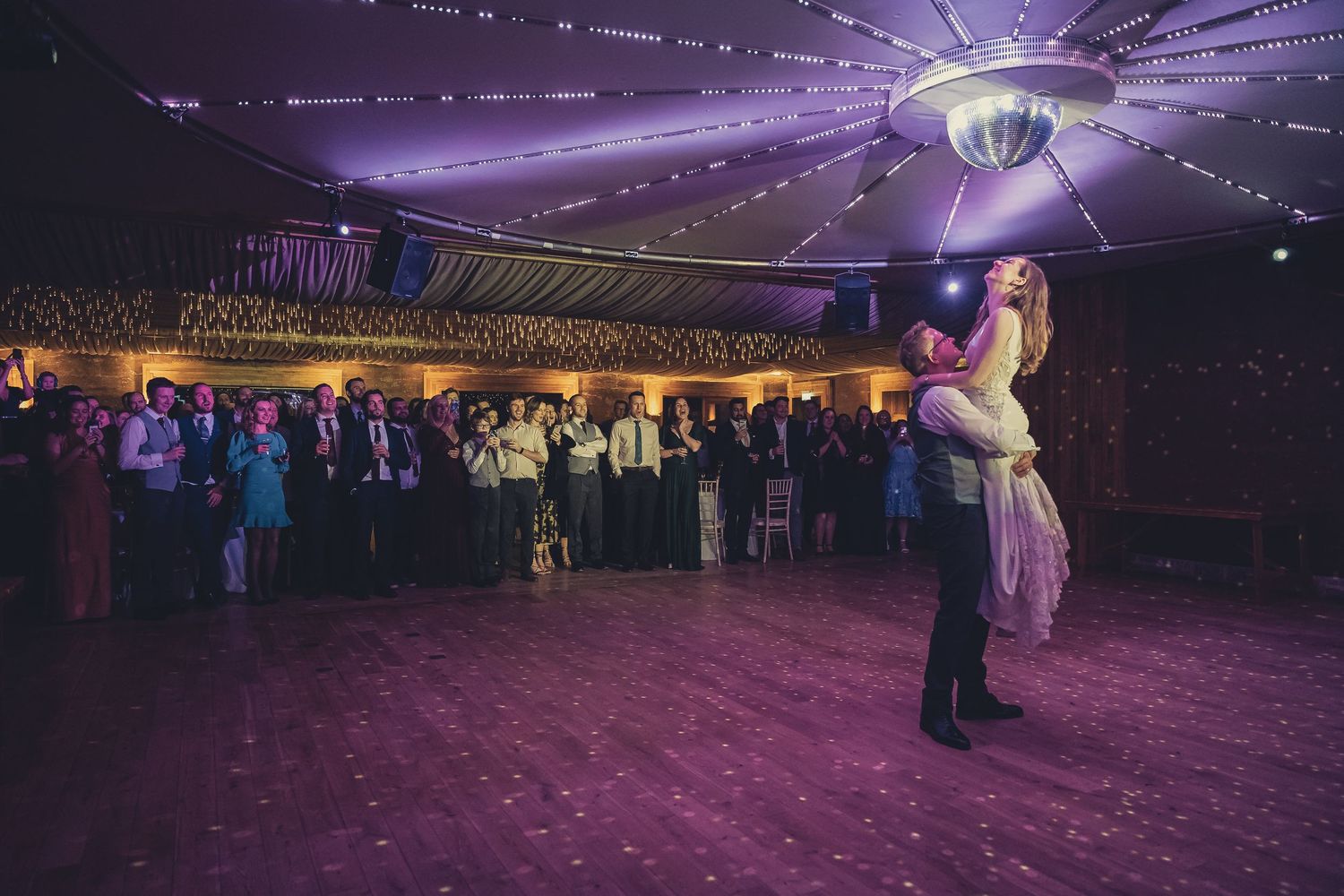 Bride lifted by groom during first dance under disco ball at elegant wedding reception.