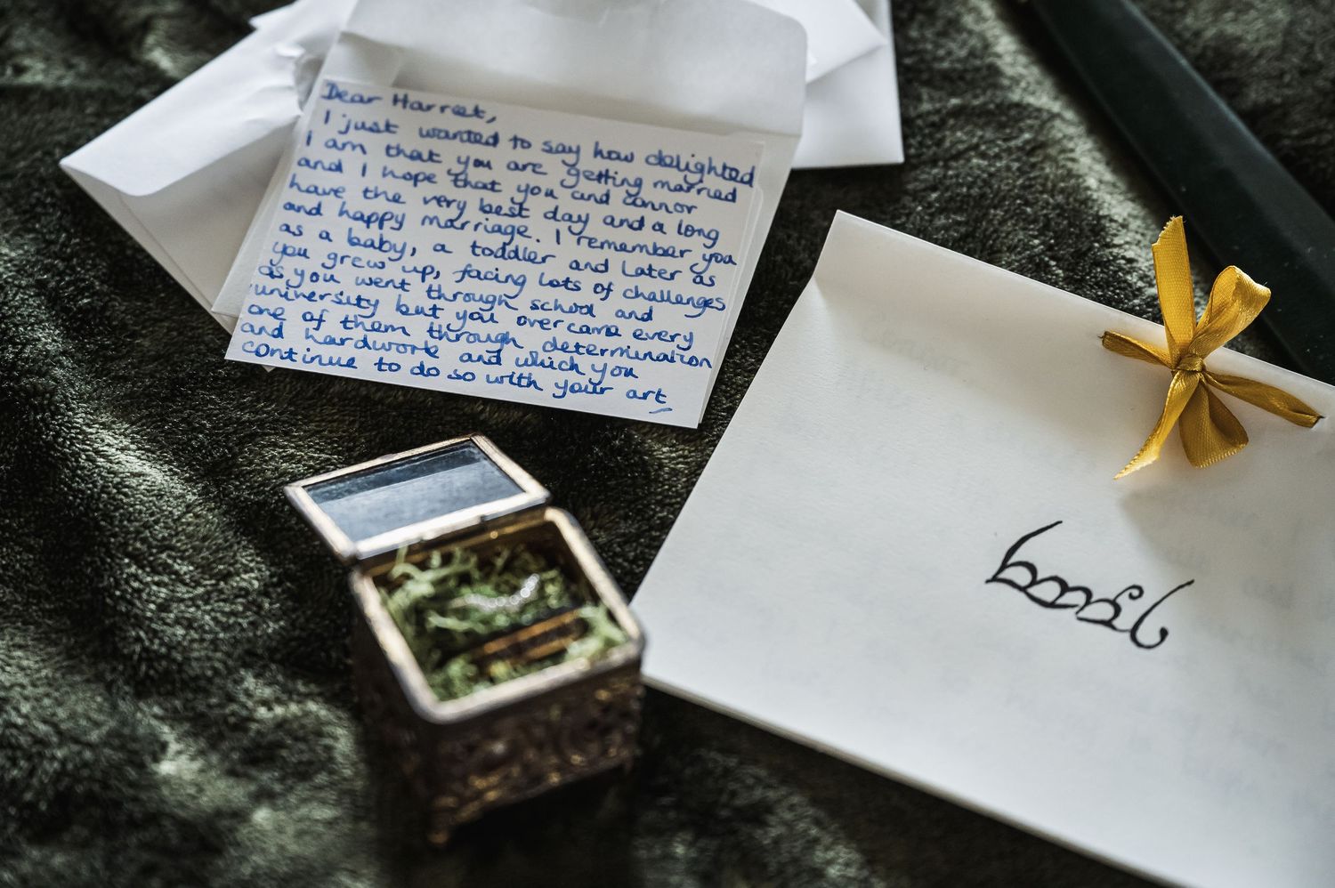 Handwritten wedding vow letter beside a small decorative box with botanical contents and an envelope on dark fabric.