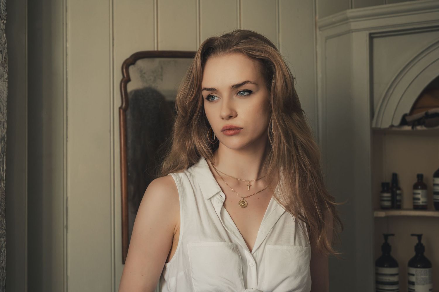 Young woman with long brown hair wearing a white sleeveless shirt and gold necklace poses indoors.