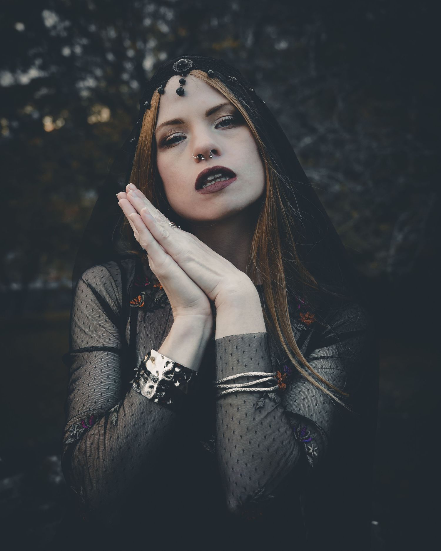Gothic woman with red hair, dark lipstick, jeweled headpiece, and studded bracelet poses with hands together.