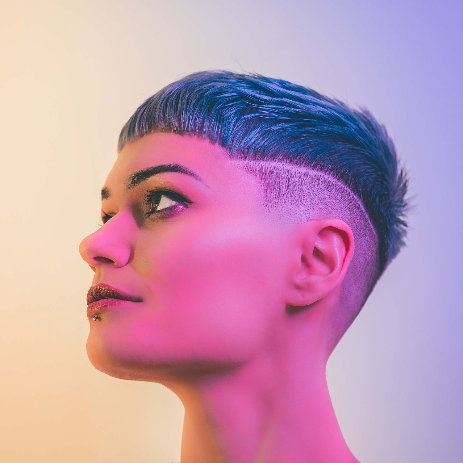 Woman with stylish undercut hairstyle photographed in profile under colorful purple and yellow lighting.