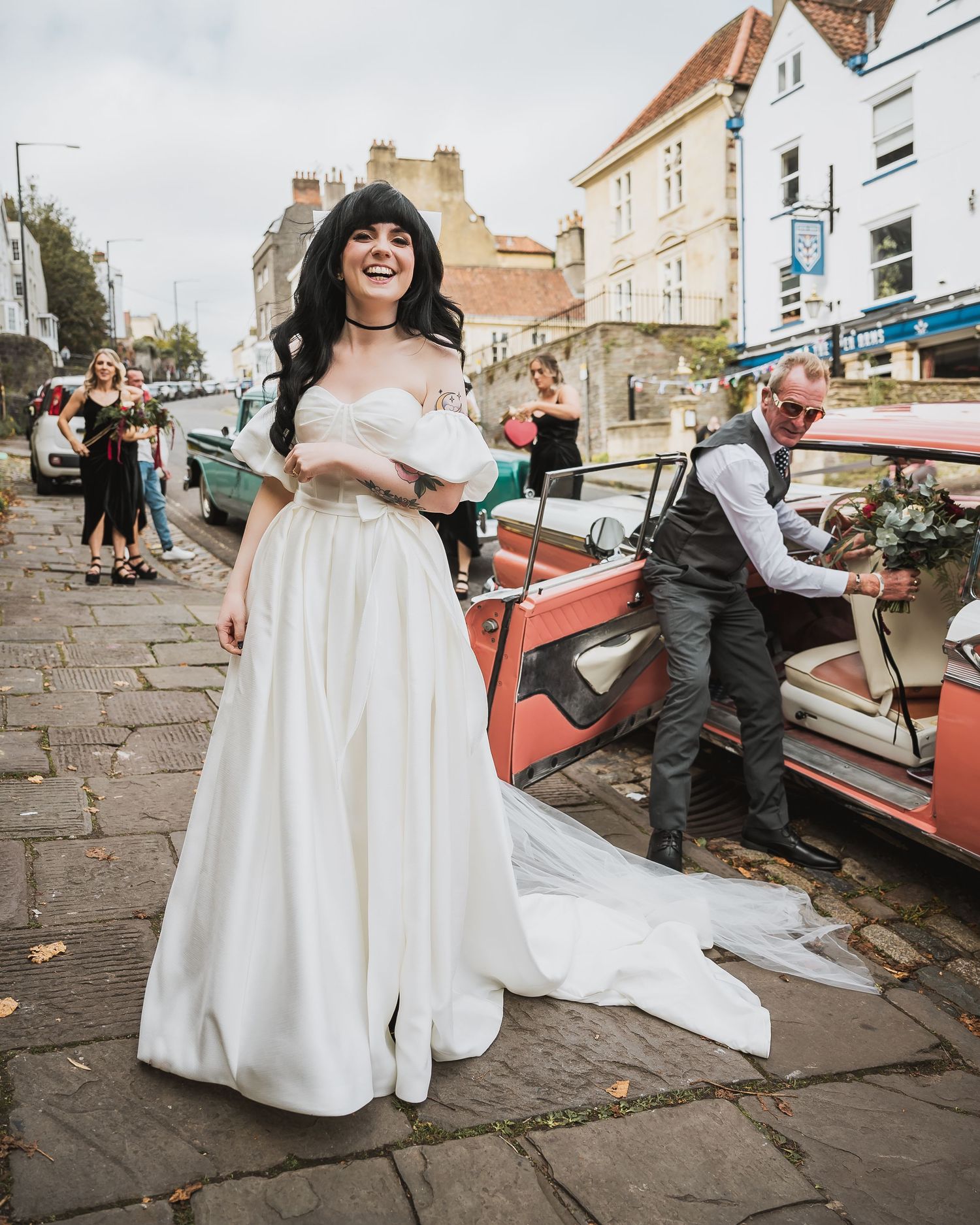 Laughing bride in off-shoulder white gown beside vintage pink car on cobblestone street.