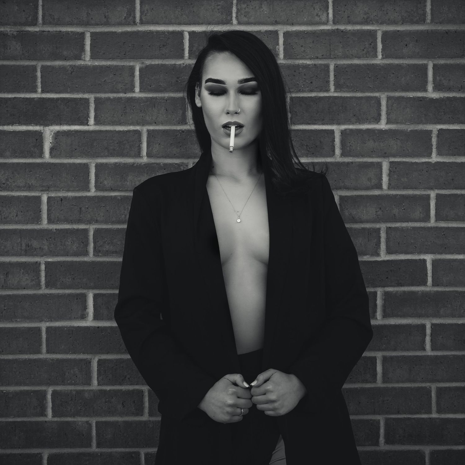 Black and white portrait of woman in open blazer smoking against brick wall.