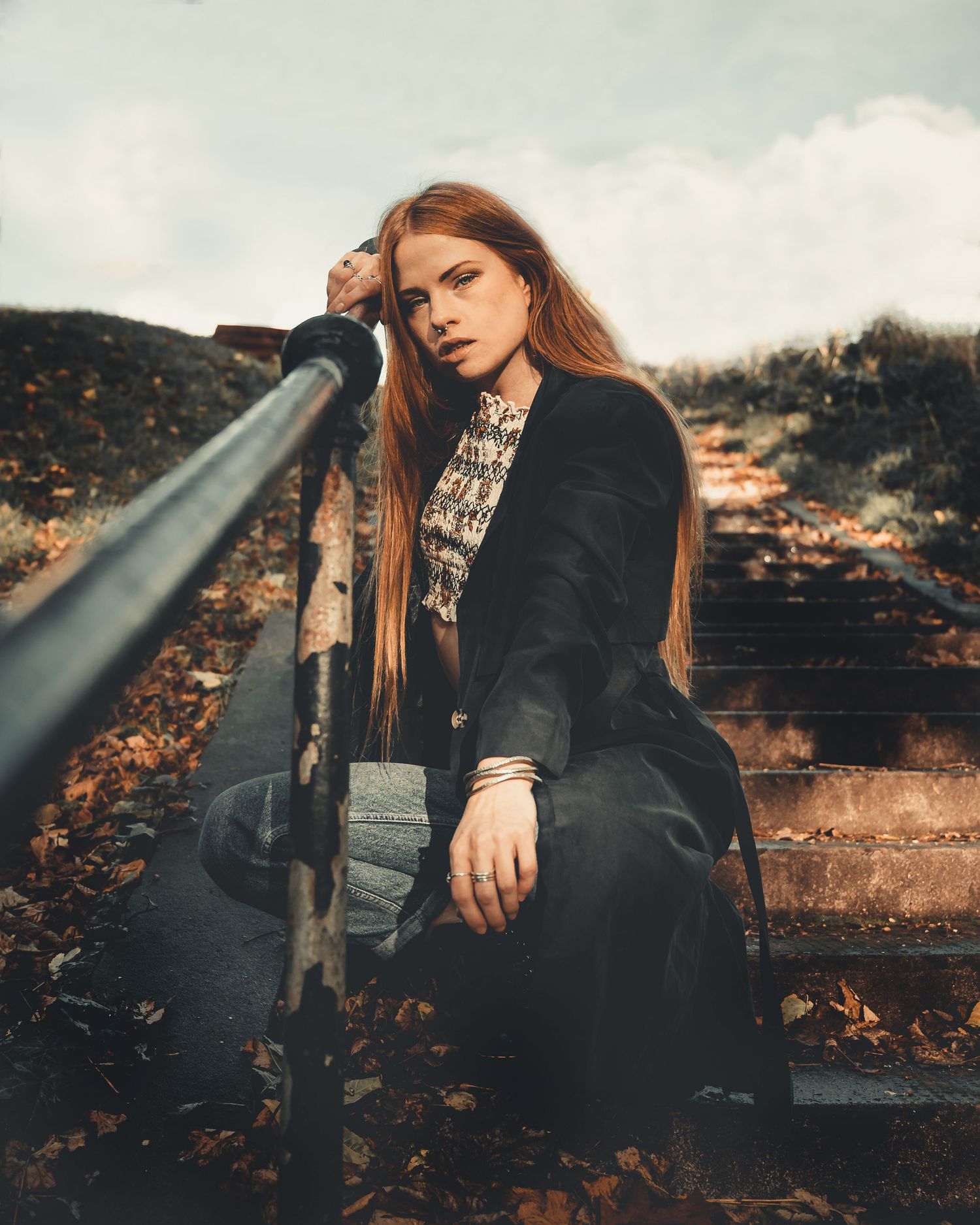 Red-haired woman in black coat sits on outdoor steps beside a metal railing among autumn leaves.