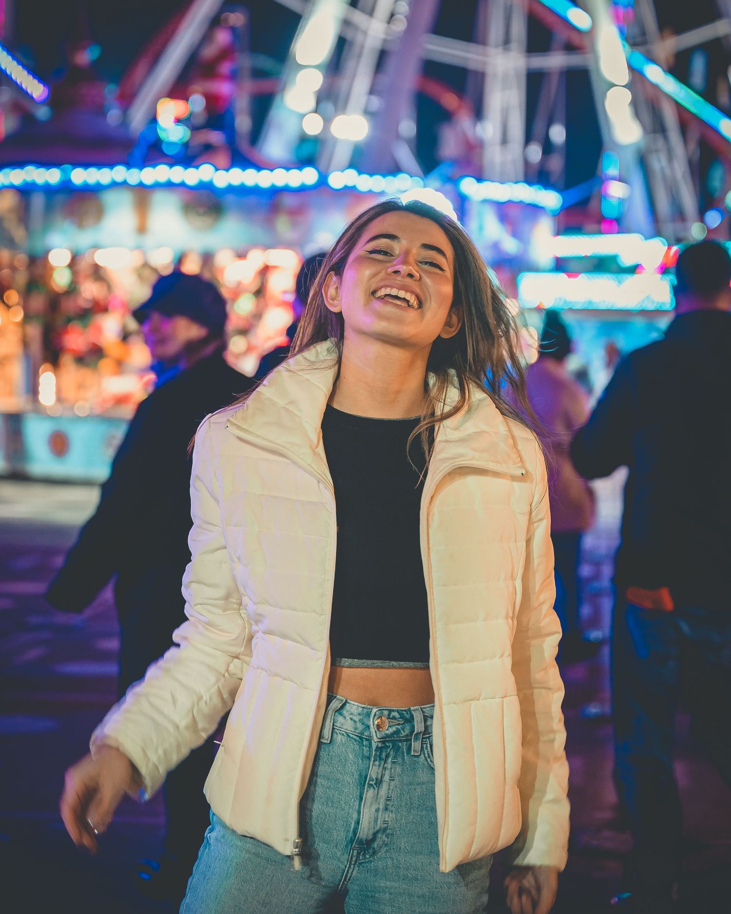 Laughing woman in white puffer jacket dancing at colorful night fairground with Ferris wheel in background.