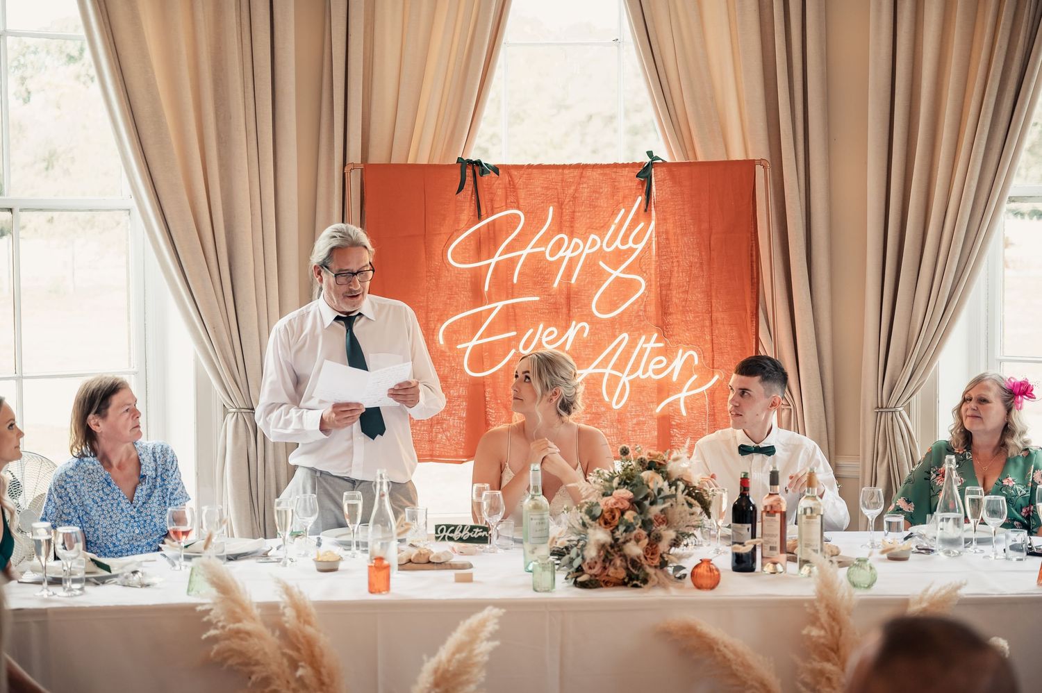 Wedding reception head table with glowing Happily Ever After neon sign backdrop during speech.