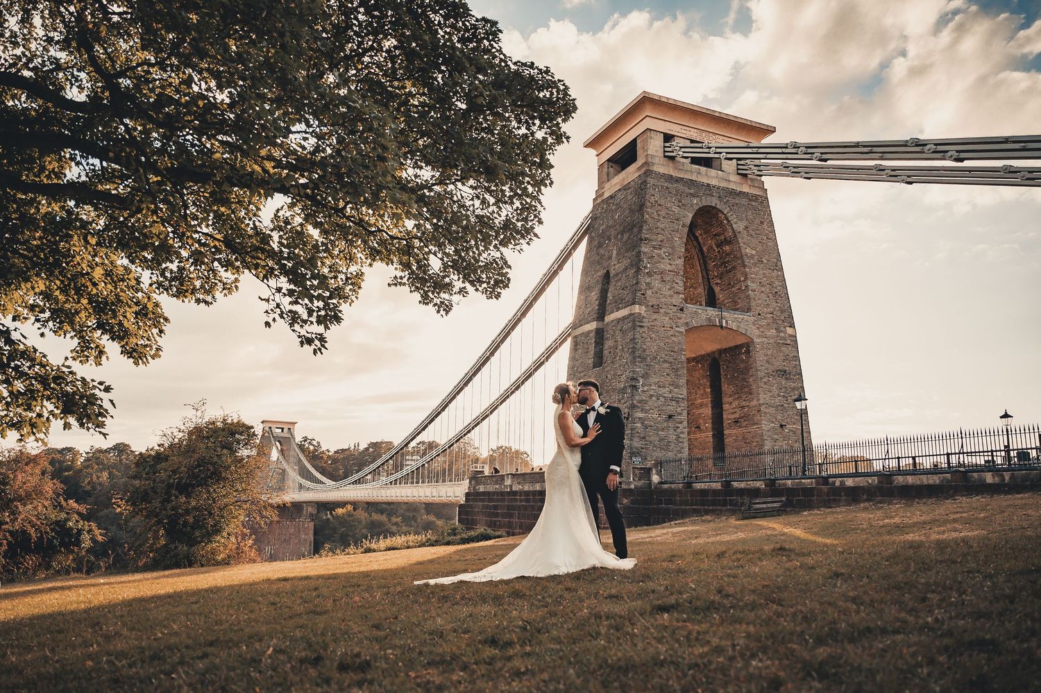 Bride and groom stand beneath the iconic Clifton Suspension Bridge tower in golden afternoon light.