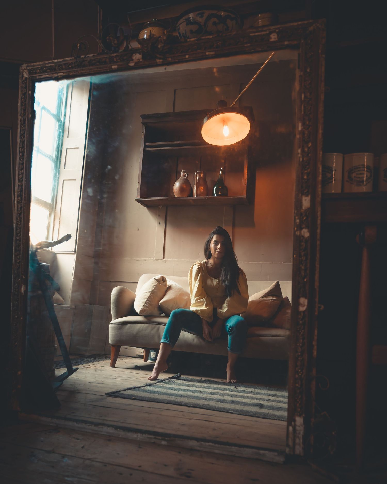 Woman in yellow top sitting on sofa reflected in ornate vintage mirror with warm lamp light.